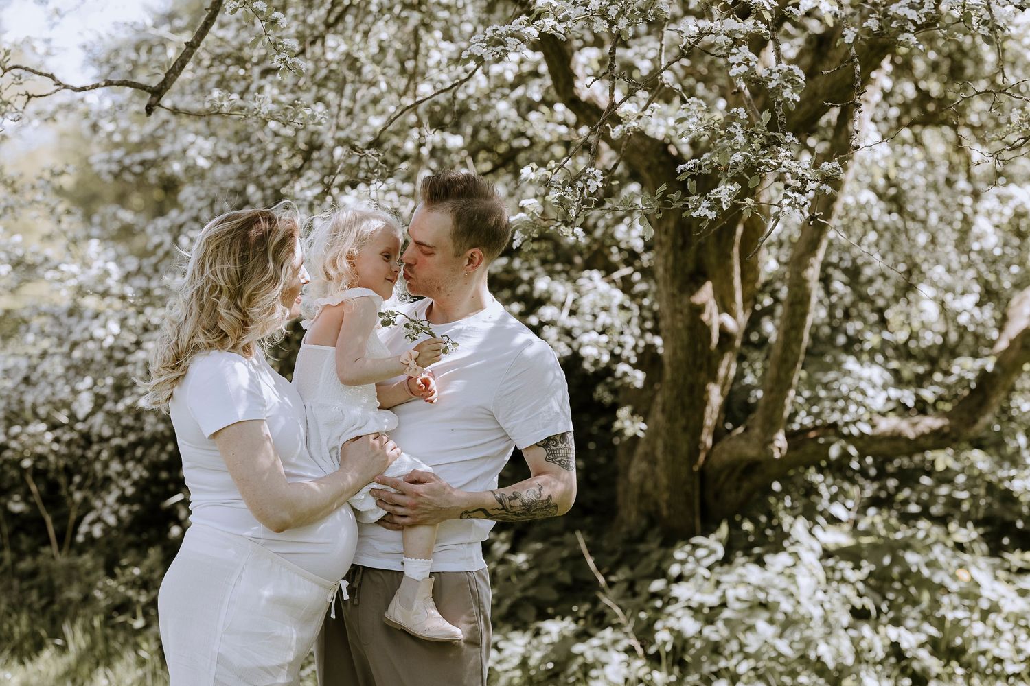 Couple in white outfits holding baby among blooming white cherry blossom trees in spring garden.