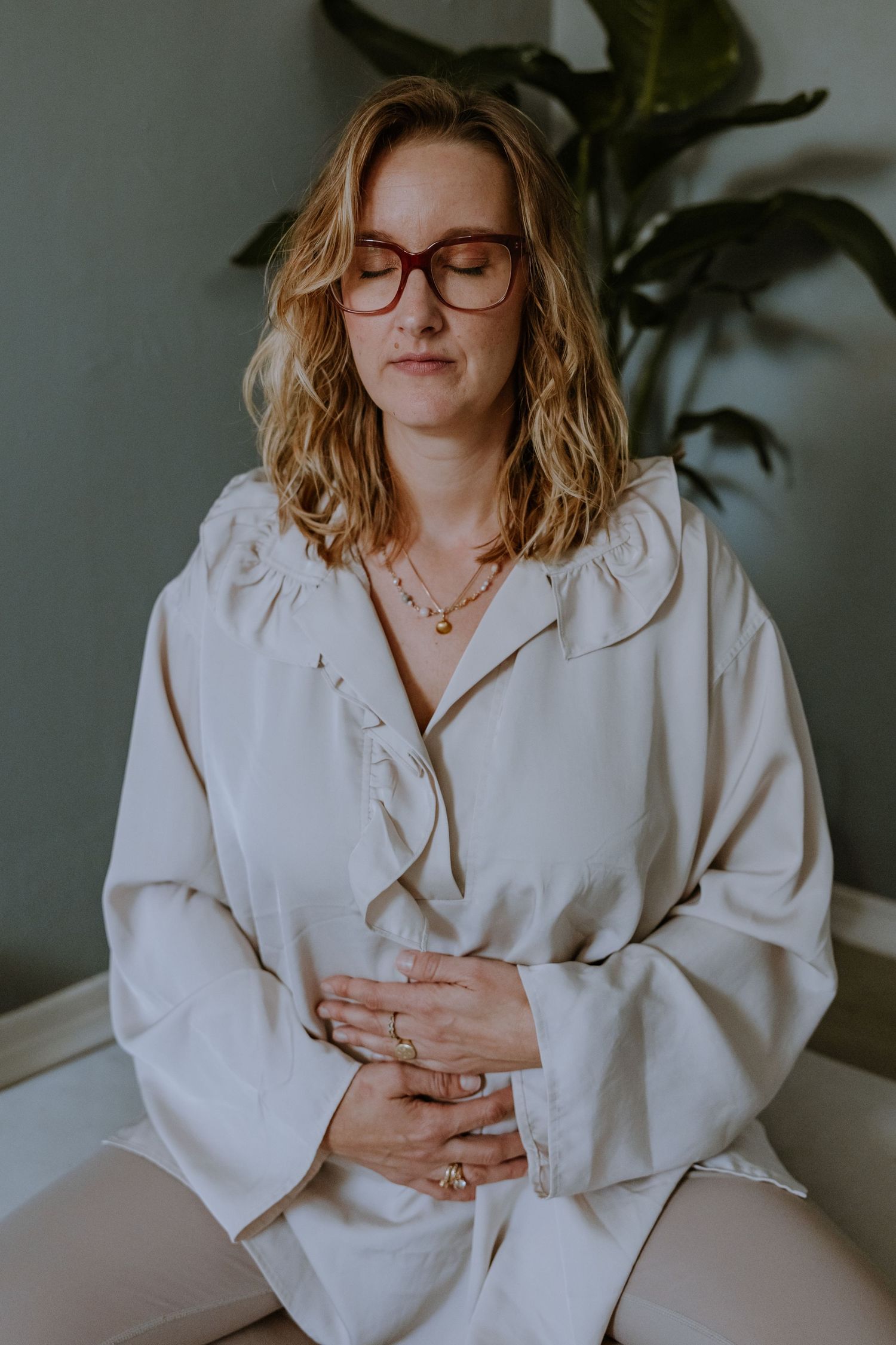 Woman in white blouse and glasses sits thoughtfully in a minimalist interior setting.