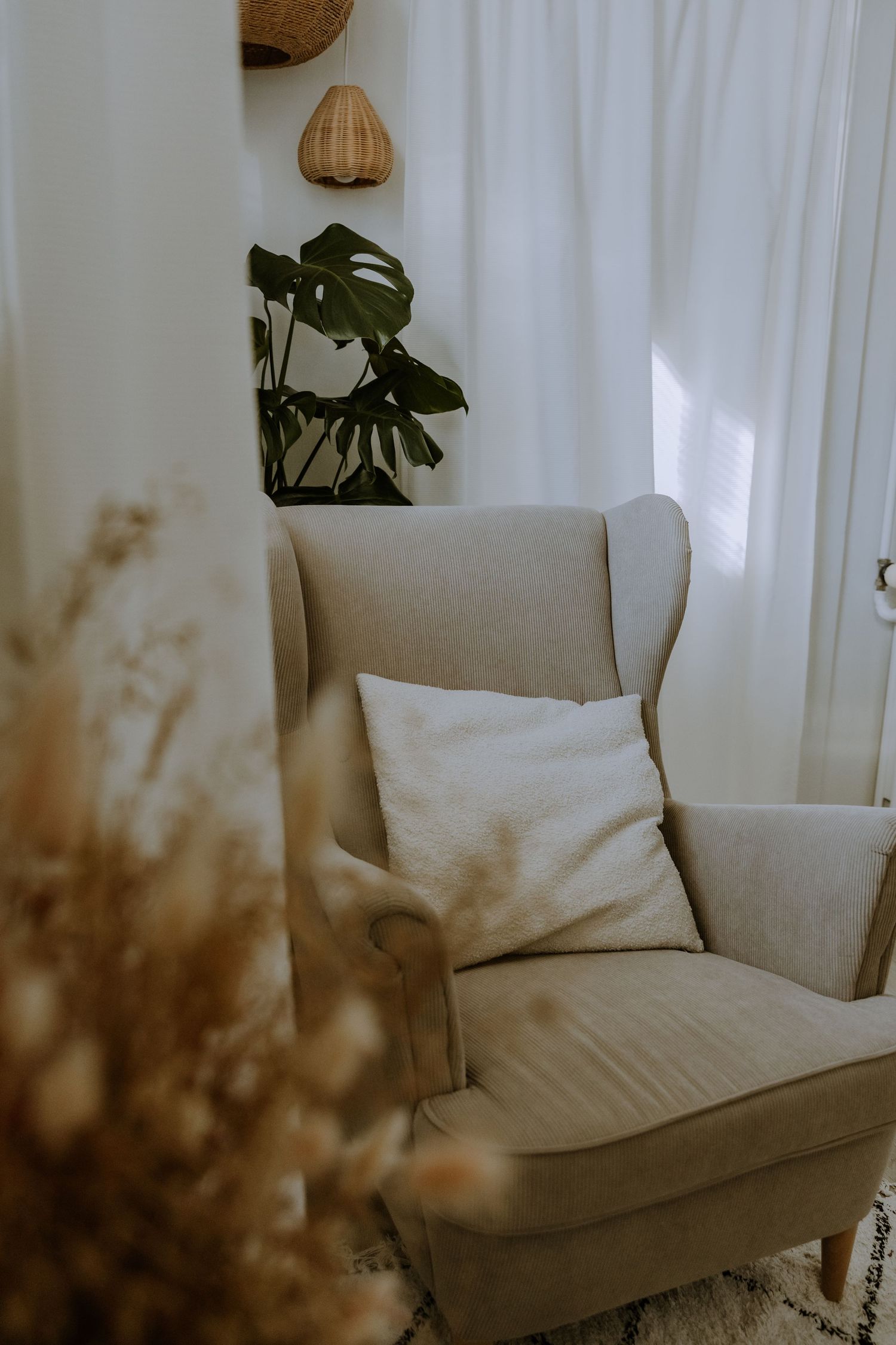 A cozy beige armchair with white pillow sits beside indoor plants and woven wall decor.