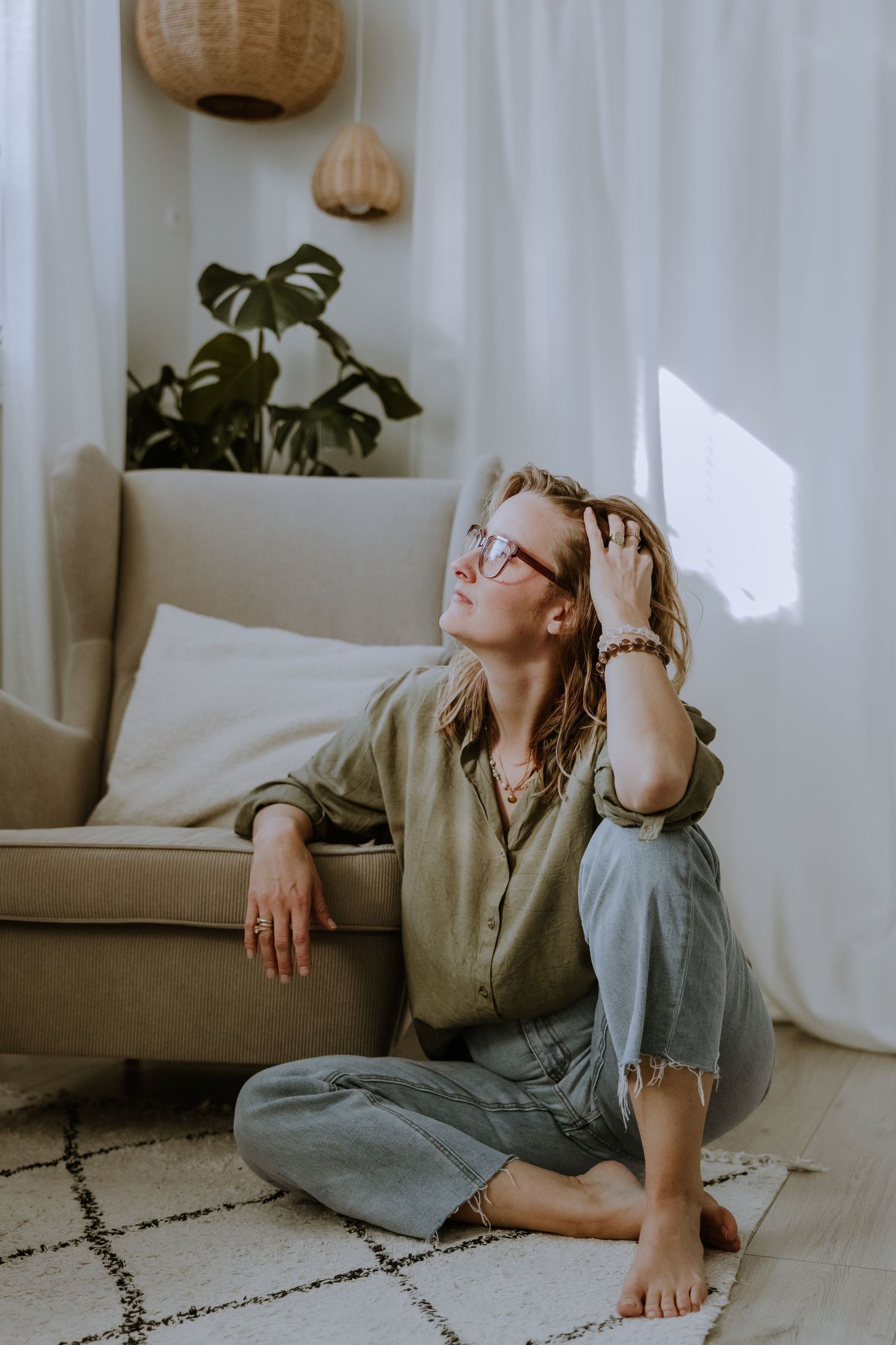 Person relaxing on floor in casual attire by cozy armchair with monstera plant and pendant lights in background.