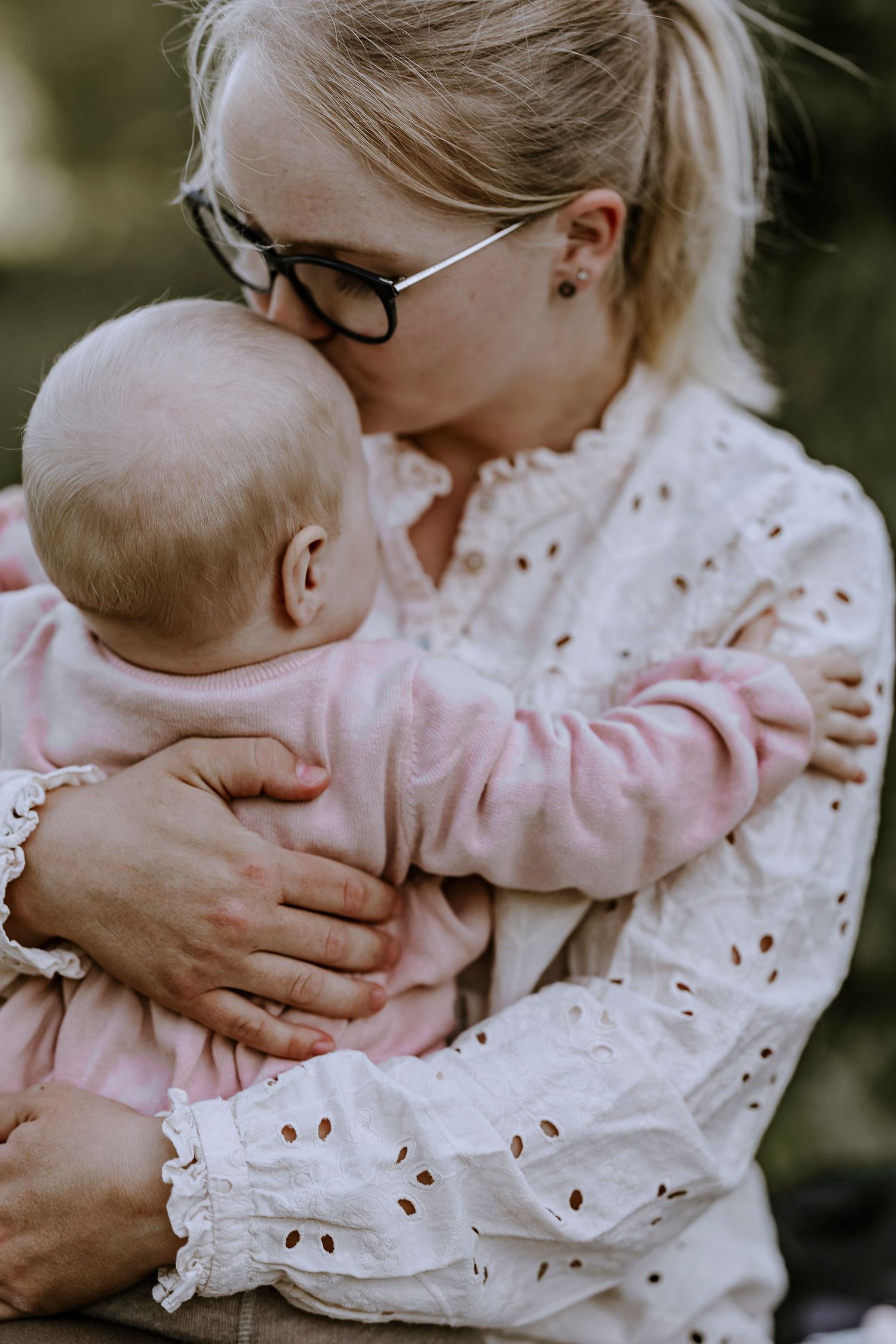 Tender moment between parent wearing eyeglasses and infant in pink clothing outdoors.