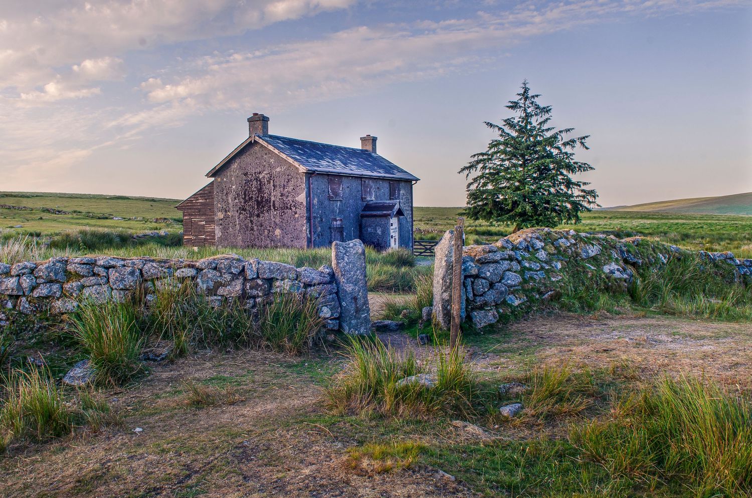 An old stone cottage stands behind a rustic wall in the moorland at dusk with rolling hills in the background.