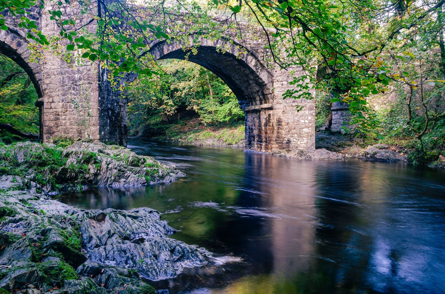 Historic stone arch holne bridge spans a tranquil river surrounded by lush greenery and rocky river dart on dartmoor