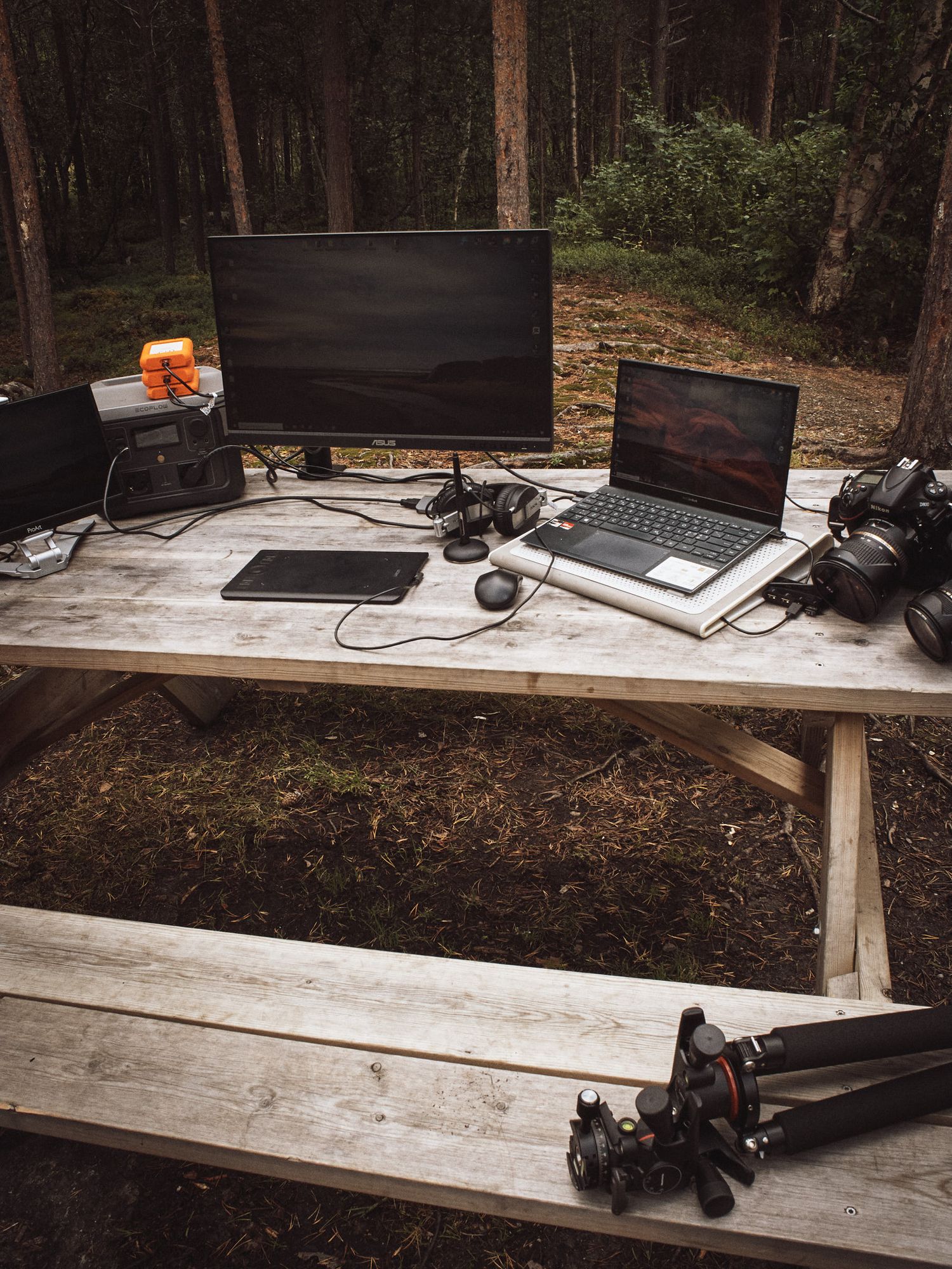 Gaming laptop and gaming peripherals arranged on a rustic wooden table setup in an outdoor forest setting.