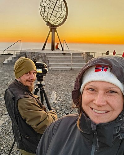 Two photographers capture sunset at Nordkapp Globe Monument in northern Norway against an orange sky.
