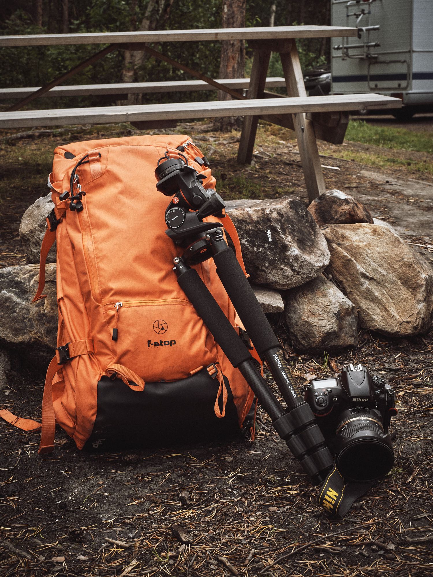 An orange F-Stop camera backpack with tripod lying on ground against rocks and leaves in outdoor setting.