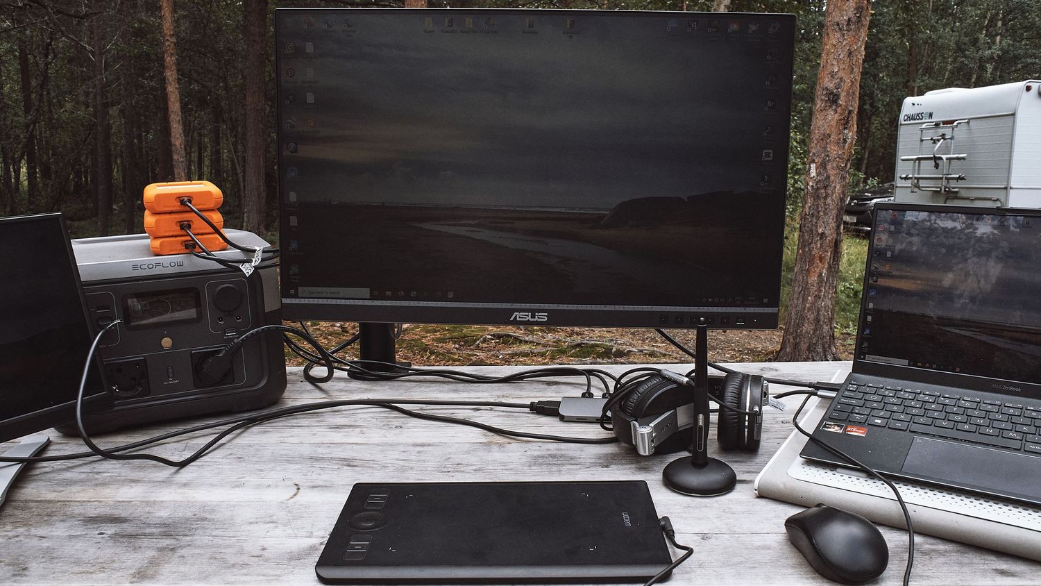 A laptop workspace setup on wooden desk with headphones, external hard drive and speakers in dark environment.