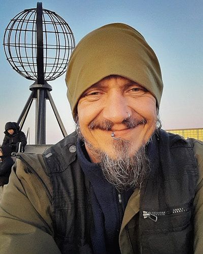 A person with a gray beanie and beard smiles while standing near a spherical wire sculpture on a sunny day.