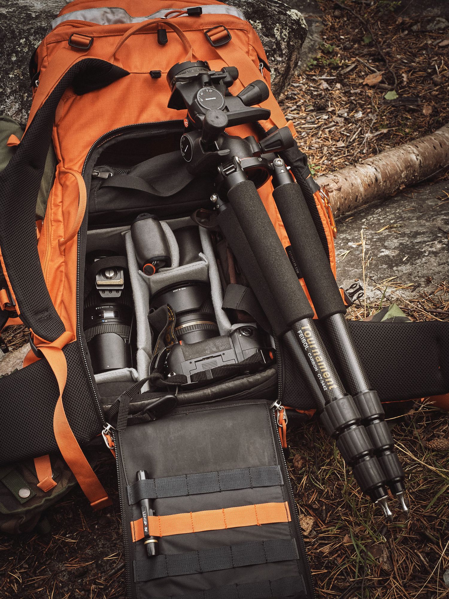An orange tactical backpack with black firearm accessories and shooting equipment displayed on forest ground.