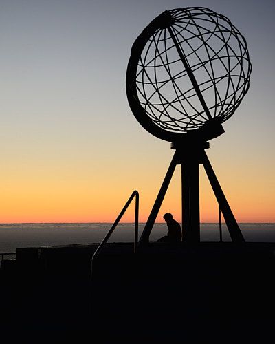 A silhouette of a person sits beneath the North Cape Globe monument against an orange sunset sky in Norway.