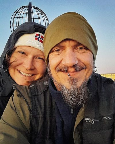Close-up outdoor selfie of two people wearing winter hats and coats, one with a Norwegian flag beanie, in warm lighting.