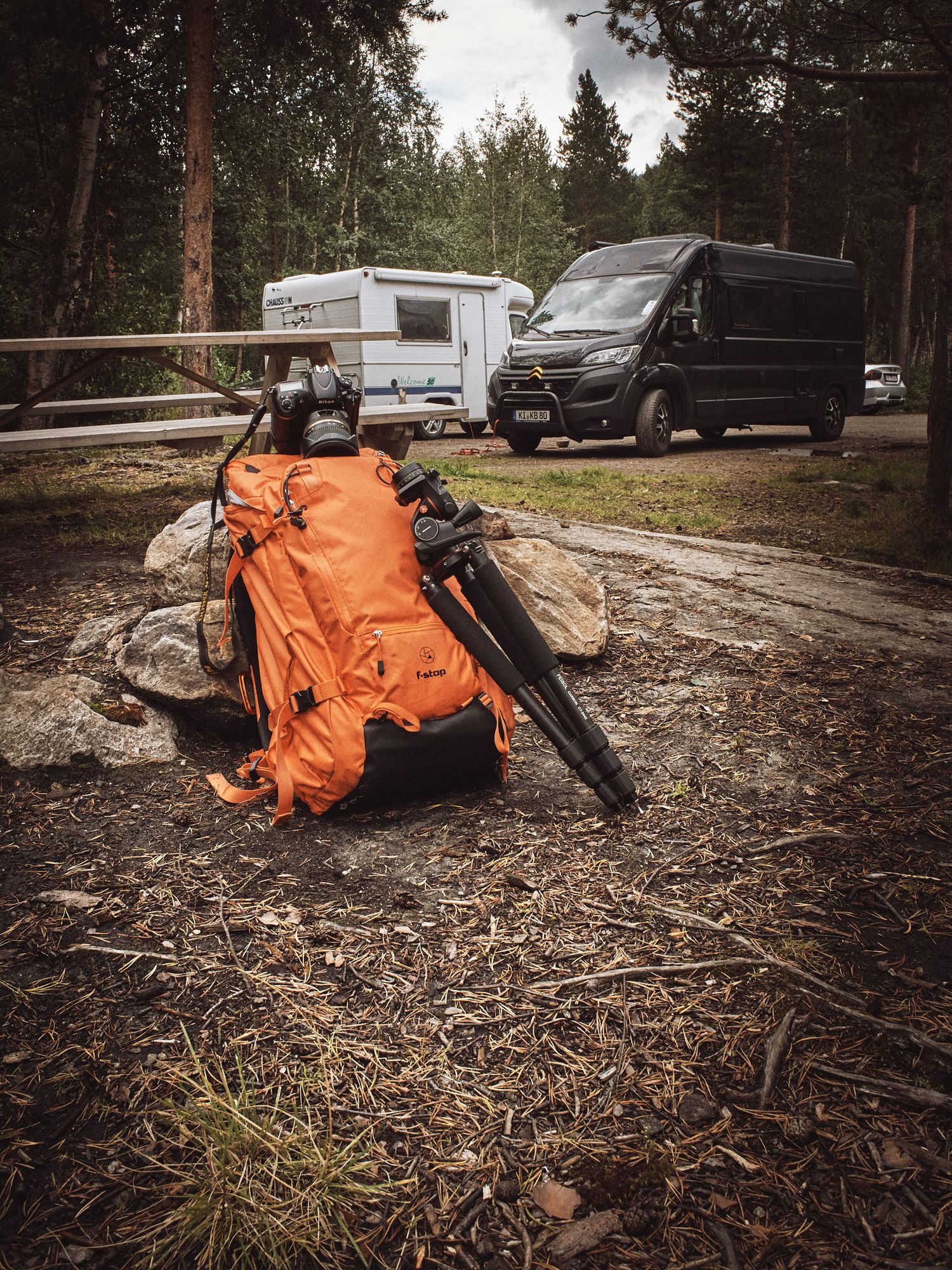 Orange camping backpack and hiking gear laid out on forest ground near RVs and vehicles in a wooded campsite.