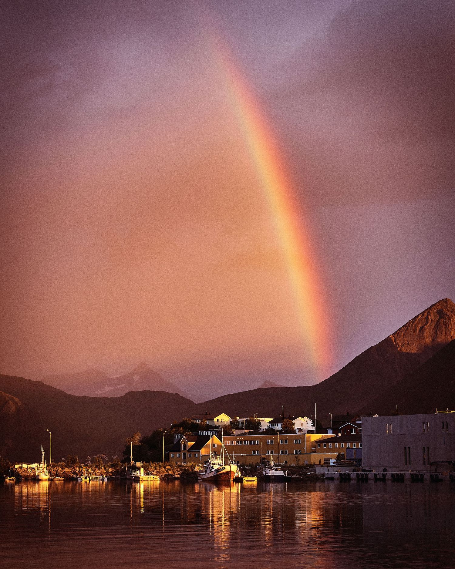 Rainbow arches over waterfront houses and mountains during golden hour.