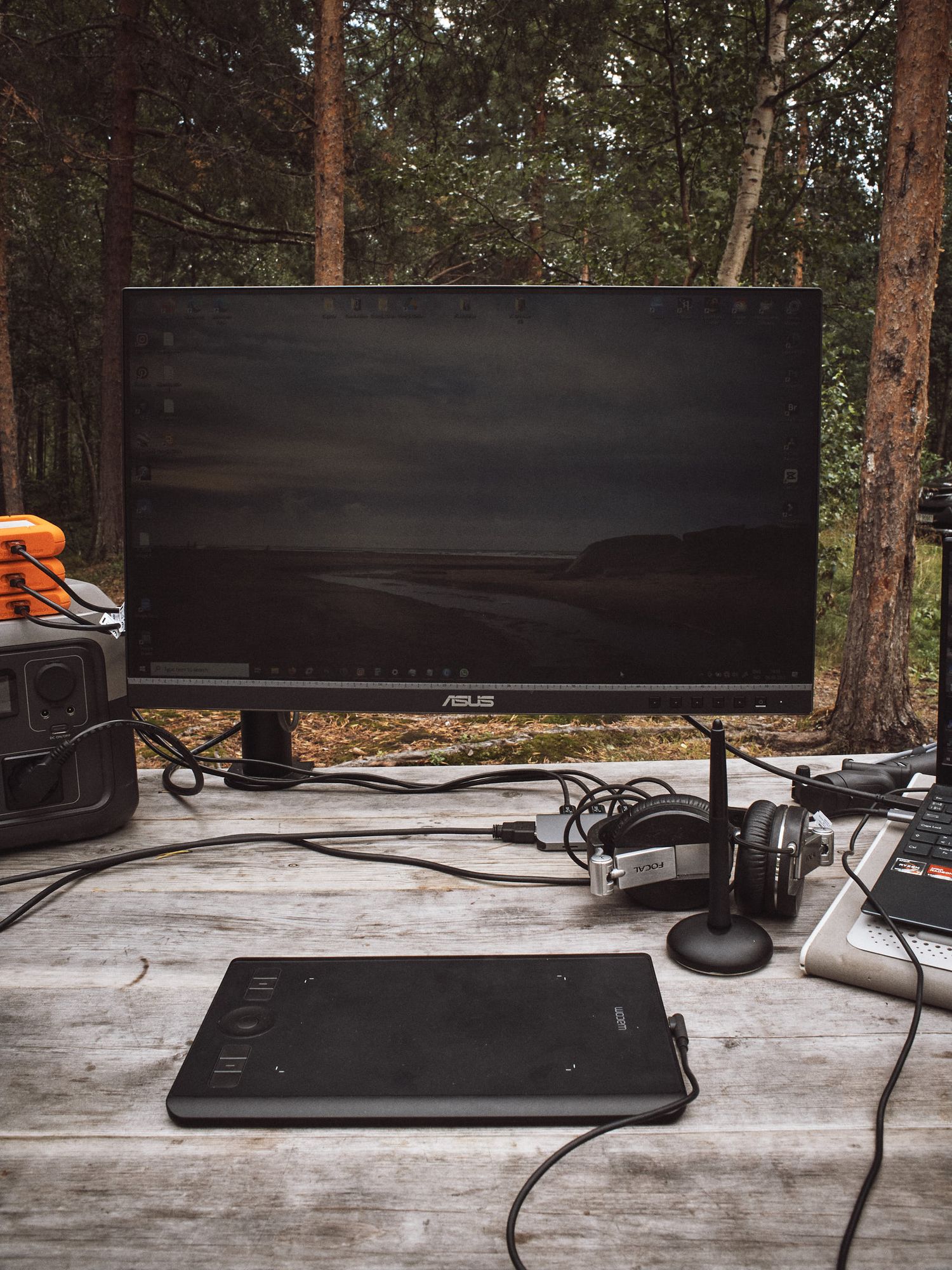 Work-from-home setup showing multiple monitors, keyboard, and mouse on gray desk surrounded by pine trees.