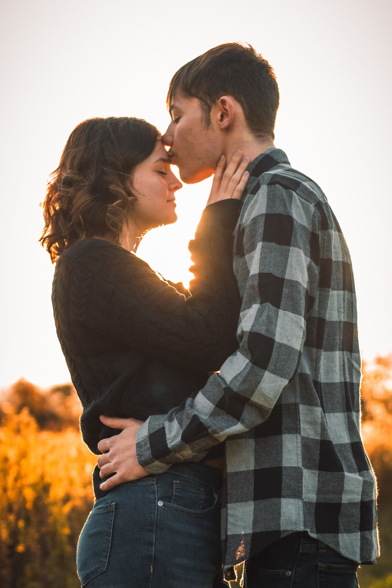Romantic couple sharing a tender moment at sunset while backlit by golden sunlight in a plaid flannel shirt and black top.