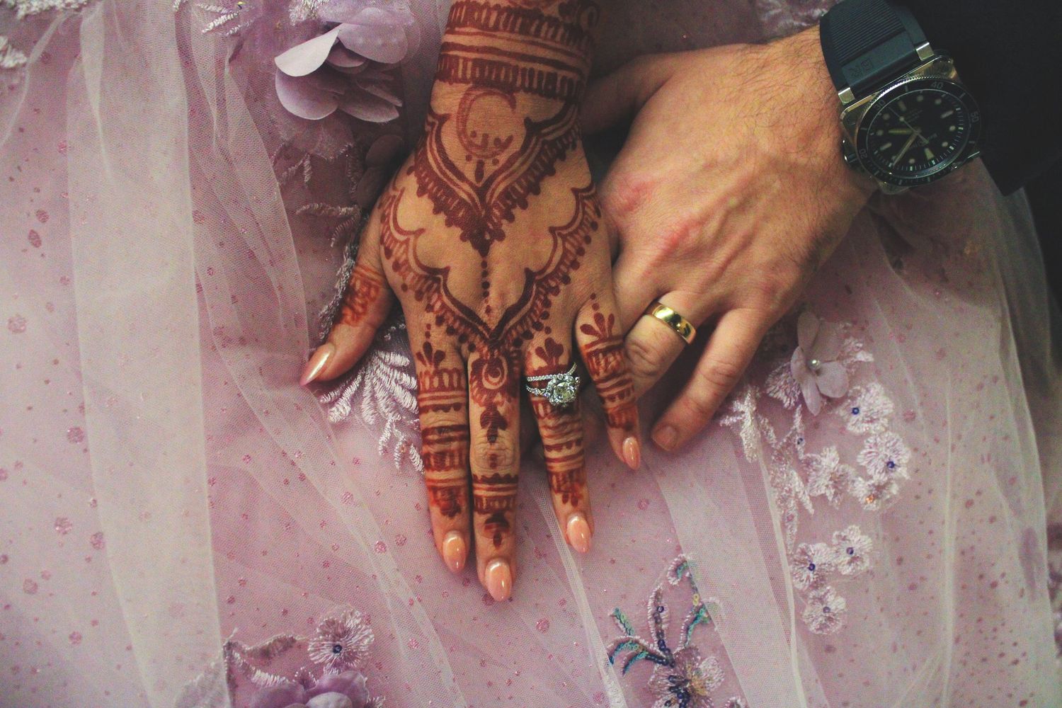 Detailed henna design adorning a hand wearing a green bracelet and ring against pink fabric background.