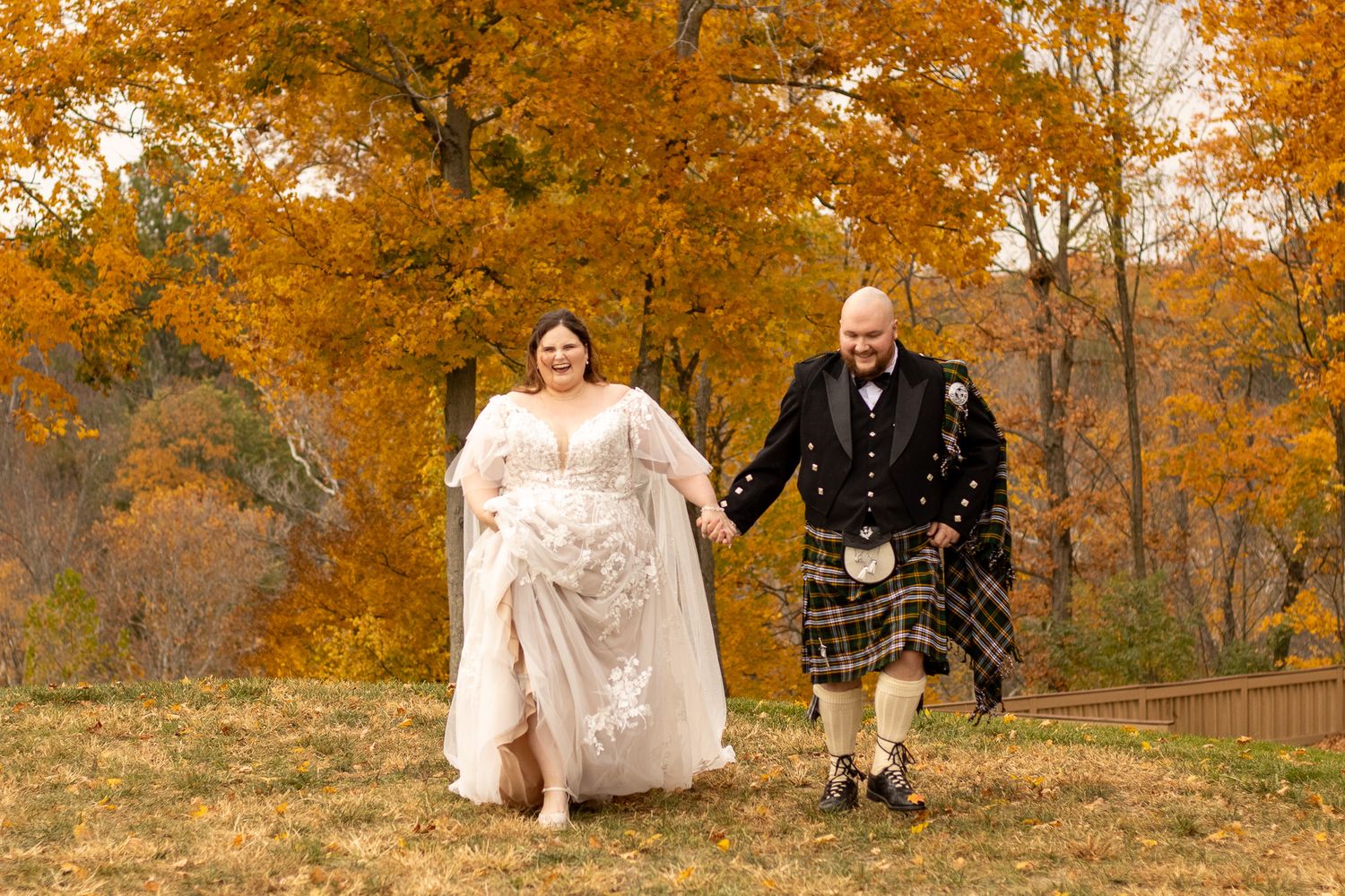 A couple holds hands while walking together in a park with vibrant autumn foliage overhead.
