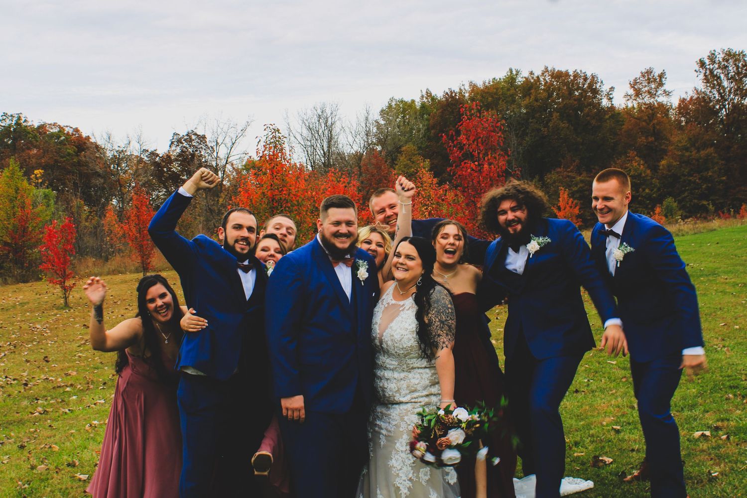 A wedding party wearing blue suits and a burgundy dress pose together on an autumn day with colorful fall foliage behind them.