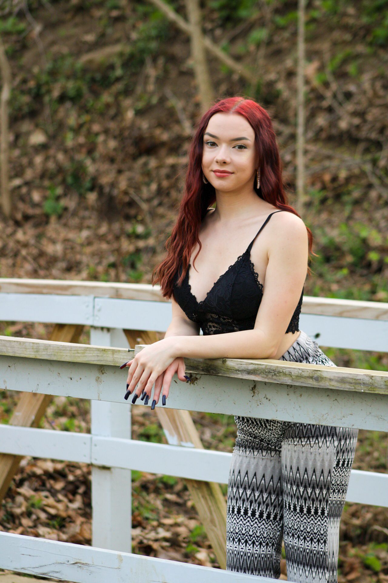 Woman with long red hair wearing black top and patterned pants poses on wooden railing outdoors.