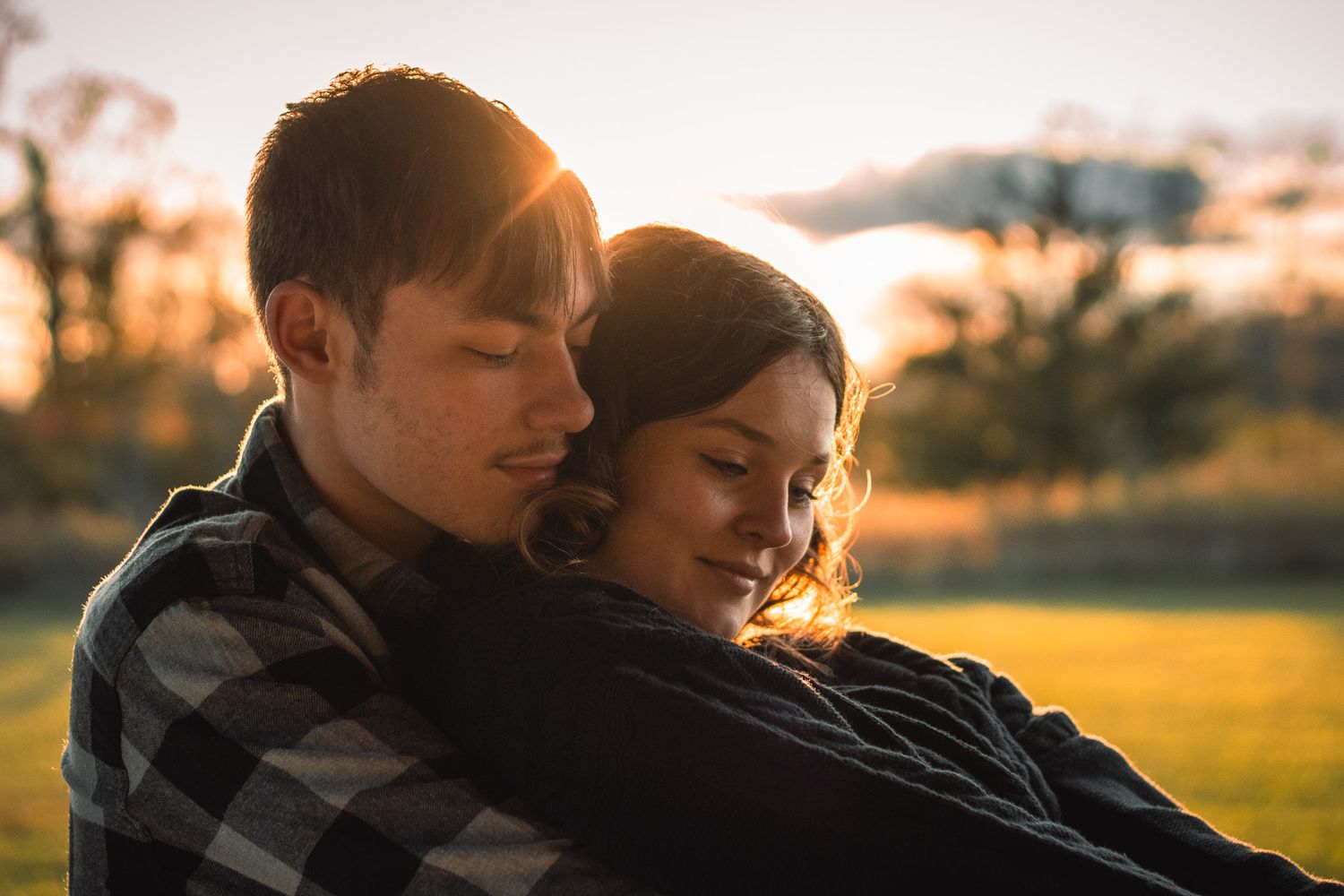 A couple shares an intimate moment in a warm golden sunset light while wearing casual plaid clothing outdoors.