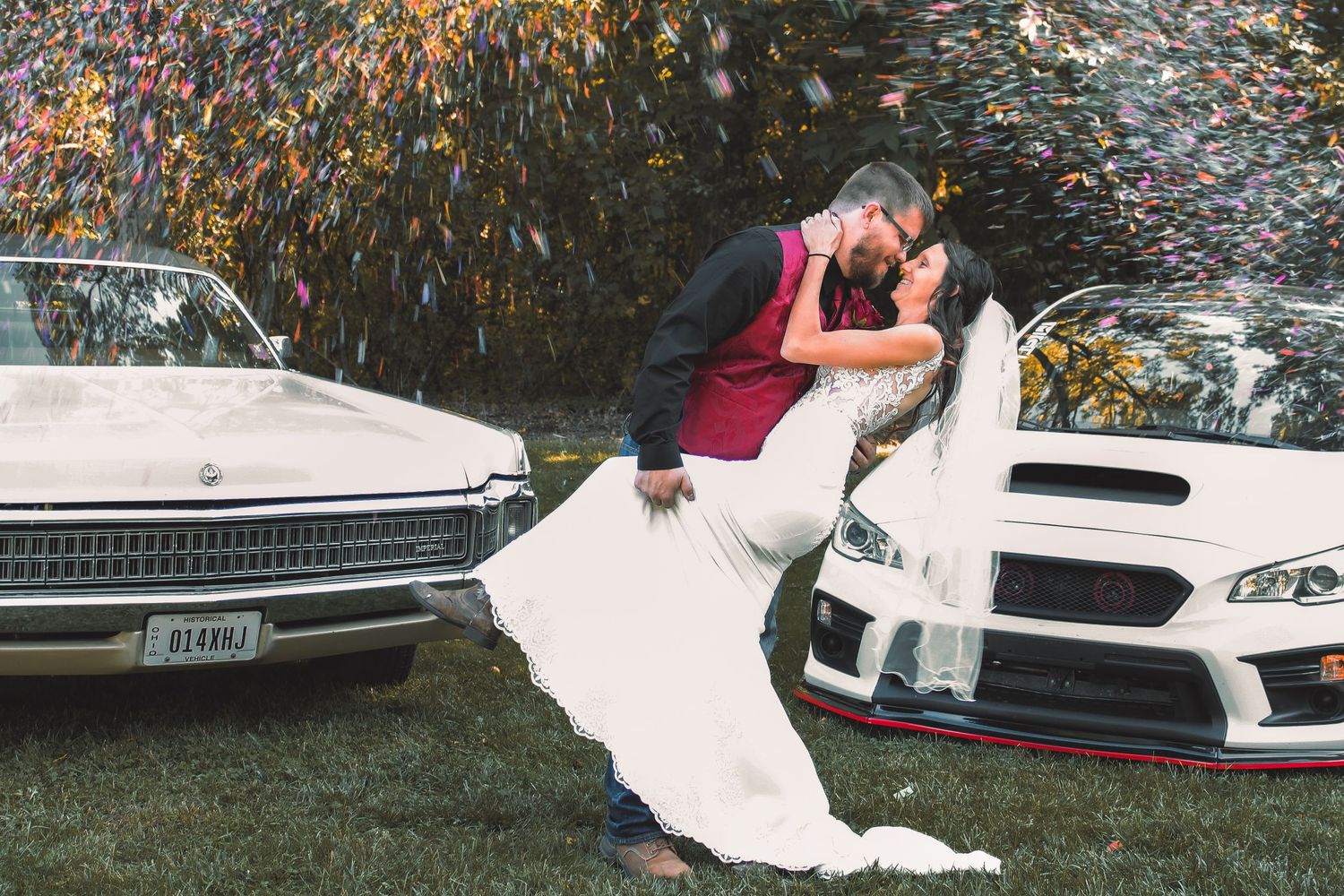 A couple embraces in a romantic dip pose between a vintage car and modern vehicle on a grassy lawn.
