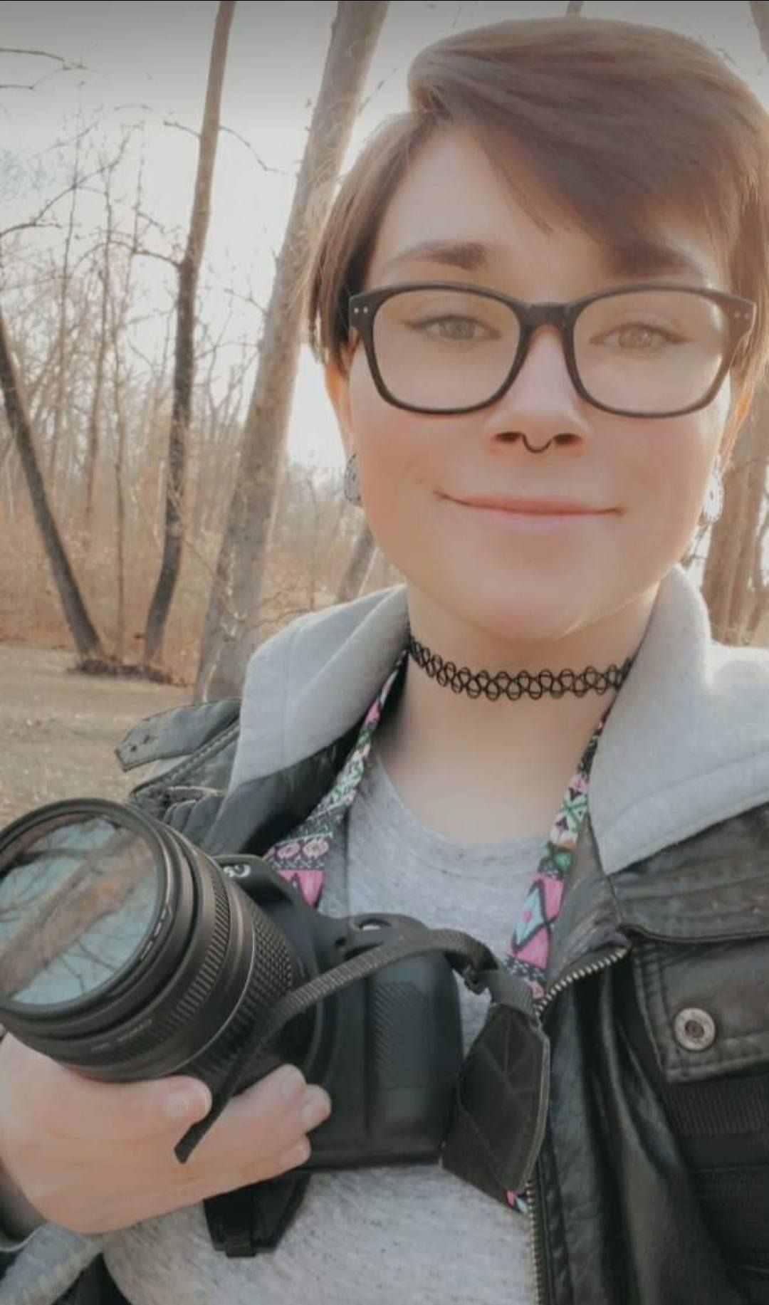 Photographer wearing glasses, choker necklace and leather jacket holds DSLR camera in wooded outdoor setting.
