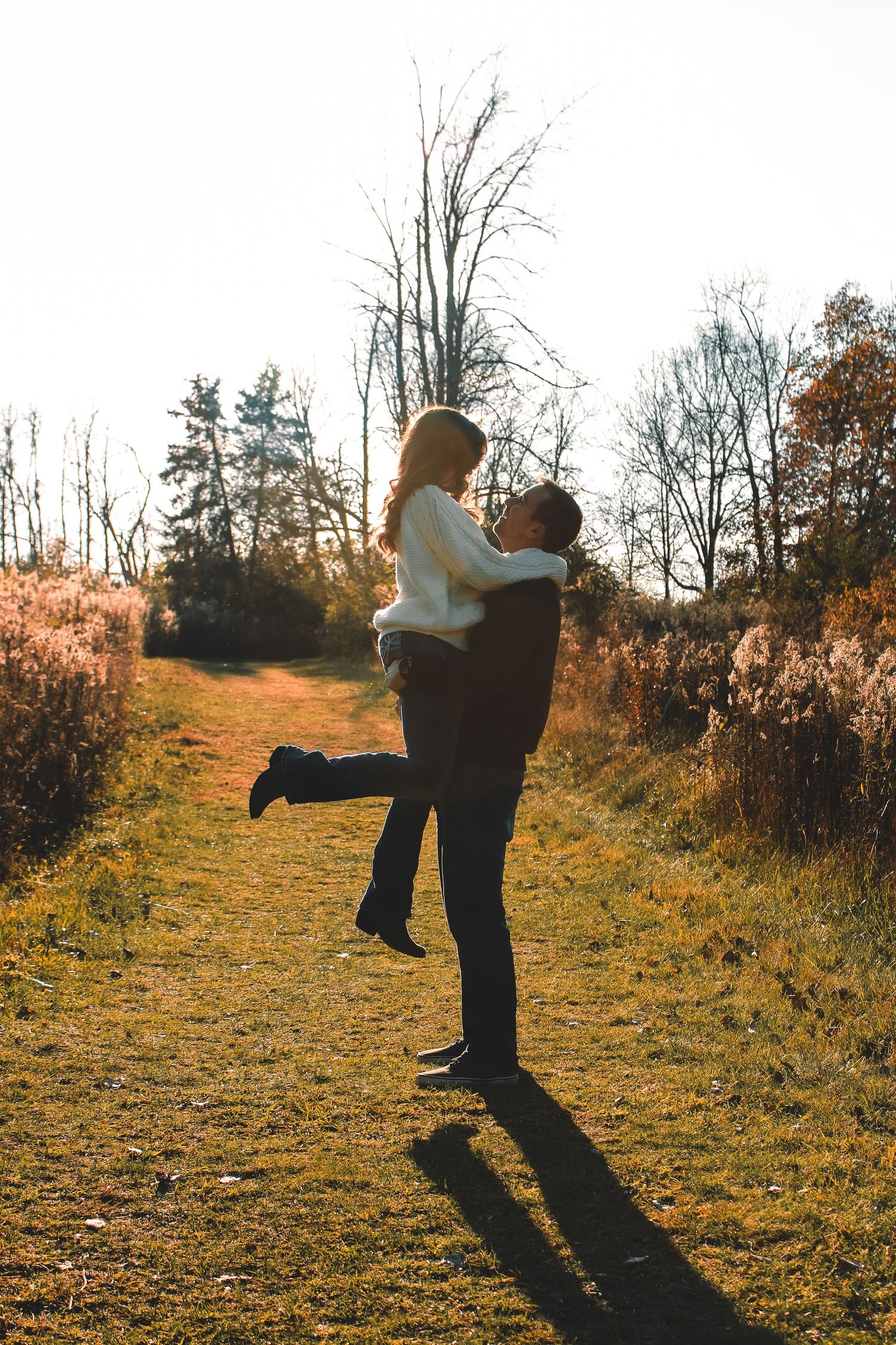 A couple embraces in a romantic autumn field during golden hour as they cast long shadows on the grassy path.