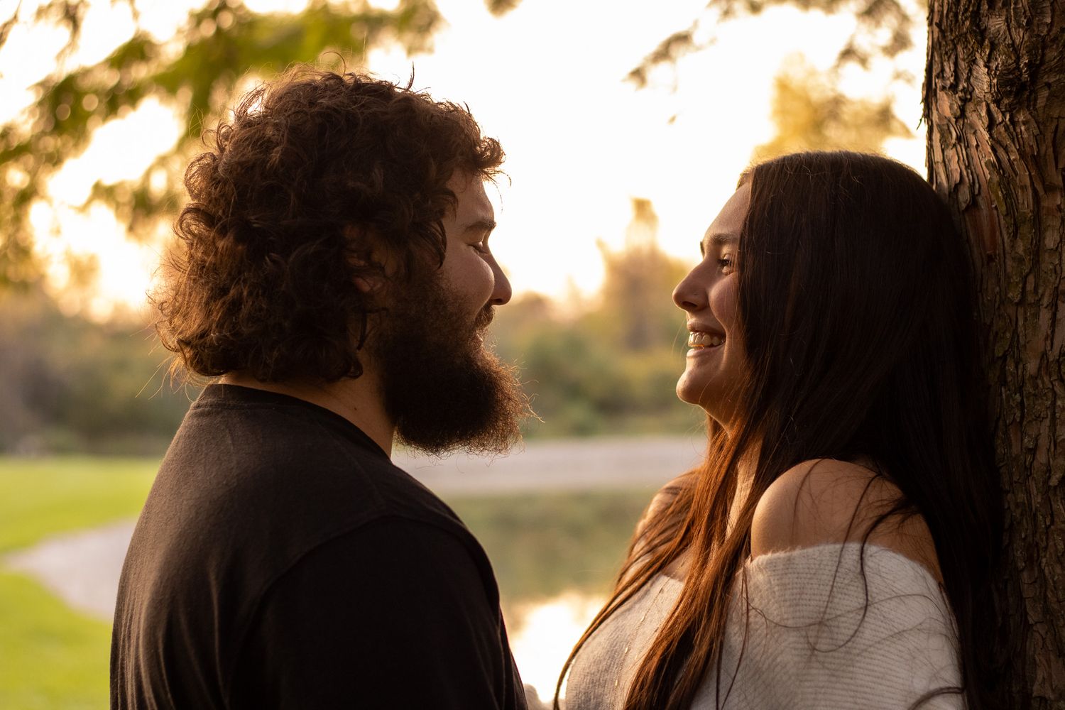 A couple smiles at each other while standing next to a tree during a warm golden sunset outdoors.