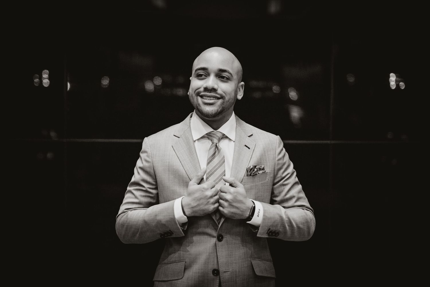 Well-dressed person in a light gray suit and tie smiles while adjusting their jacket in a black and white photo.