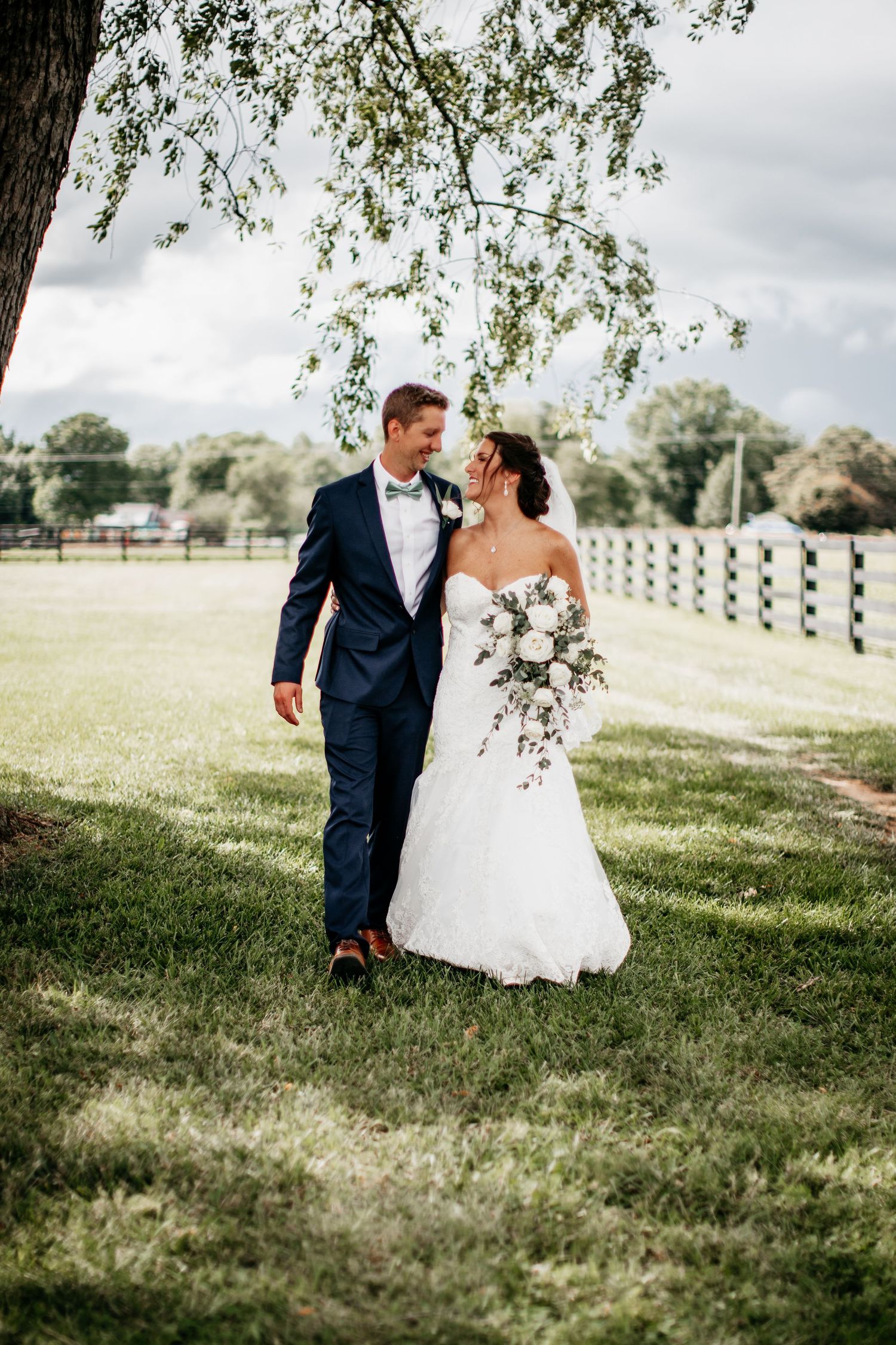 A couple walking together on a sunny day near a white fence under a willow tree in their wedding attire.