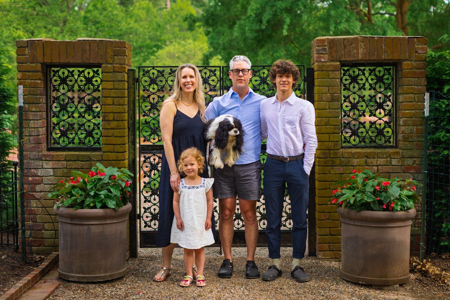 A family group poses together with their dog in front of an ornate garden gate with brick pillars and potted plants.