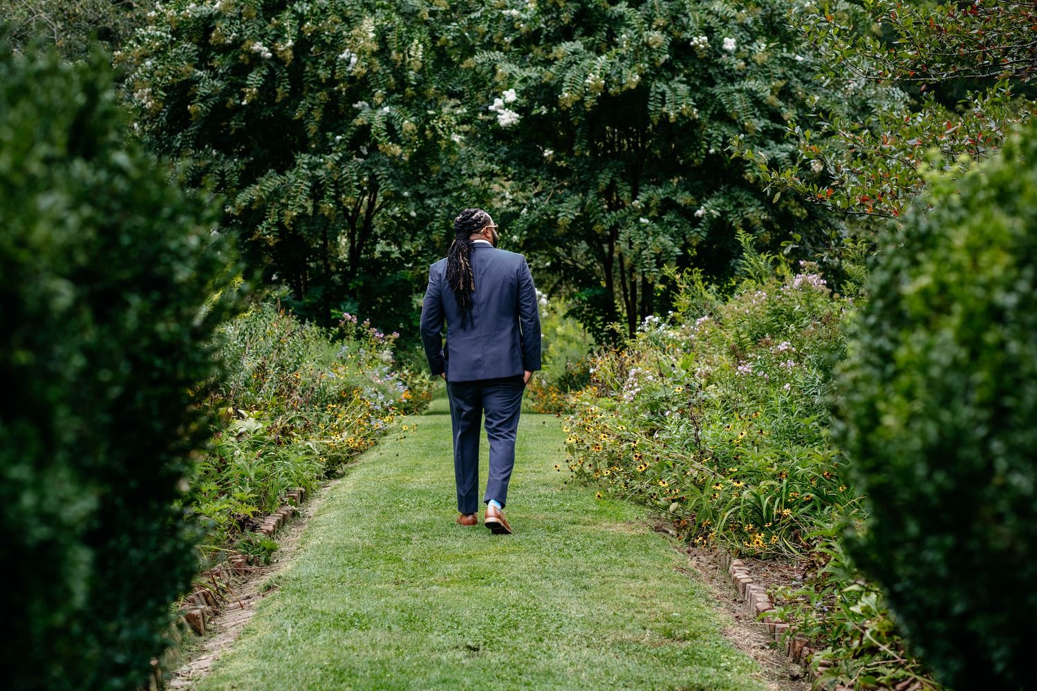 A person walks along a grassy path bordered by lush green shrubs and flowering plants in a peaceful garden setting.