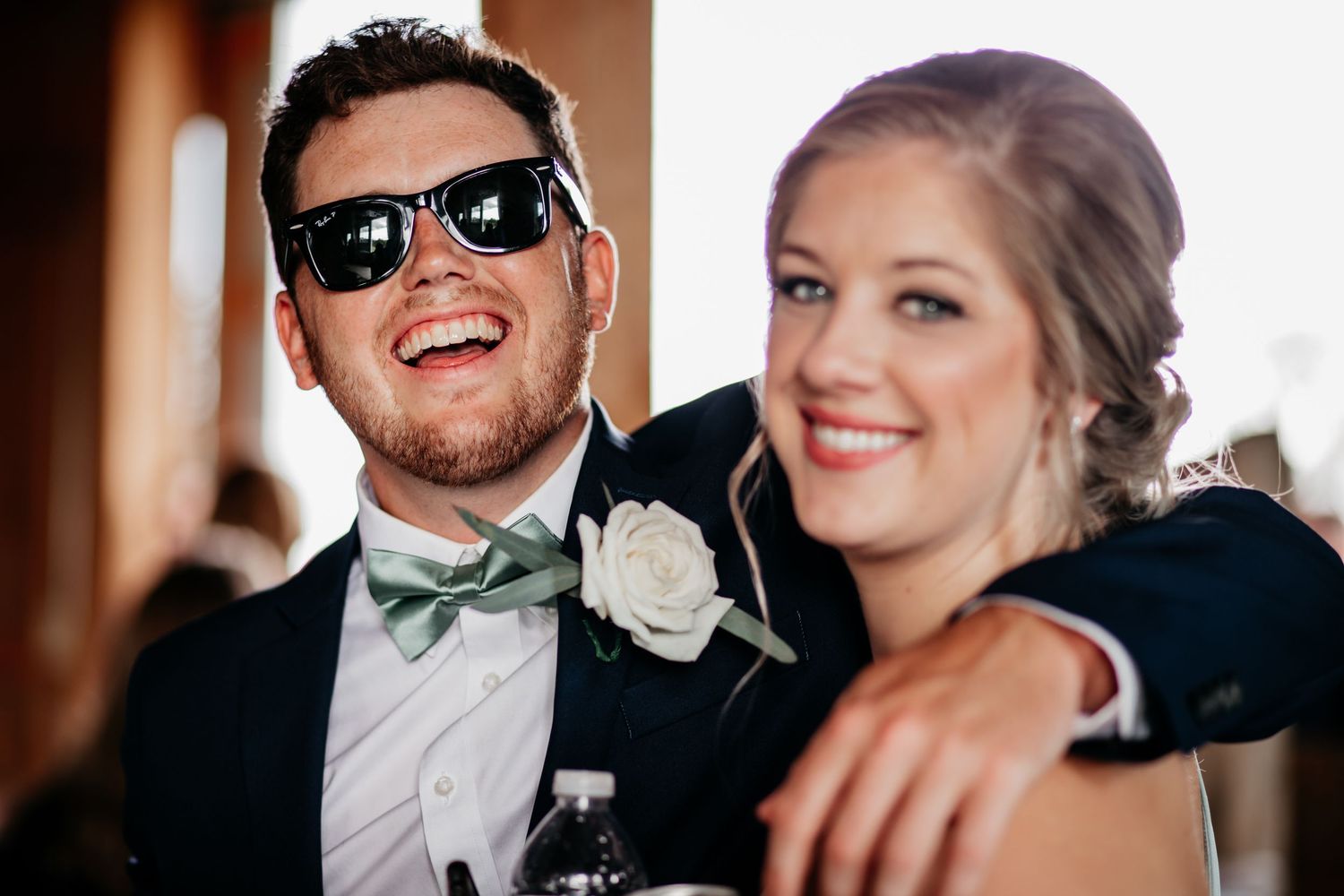 Happy couple in formal wedding attire share a joyful moment together with the groom wearing sunglasses and a bowtie.