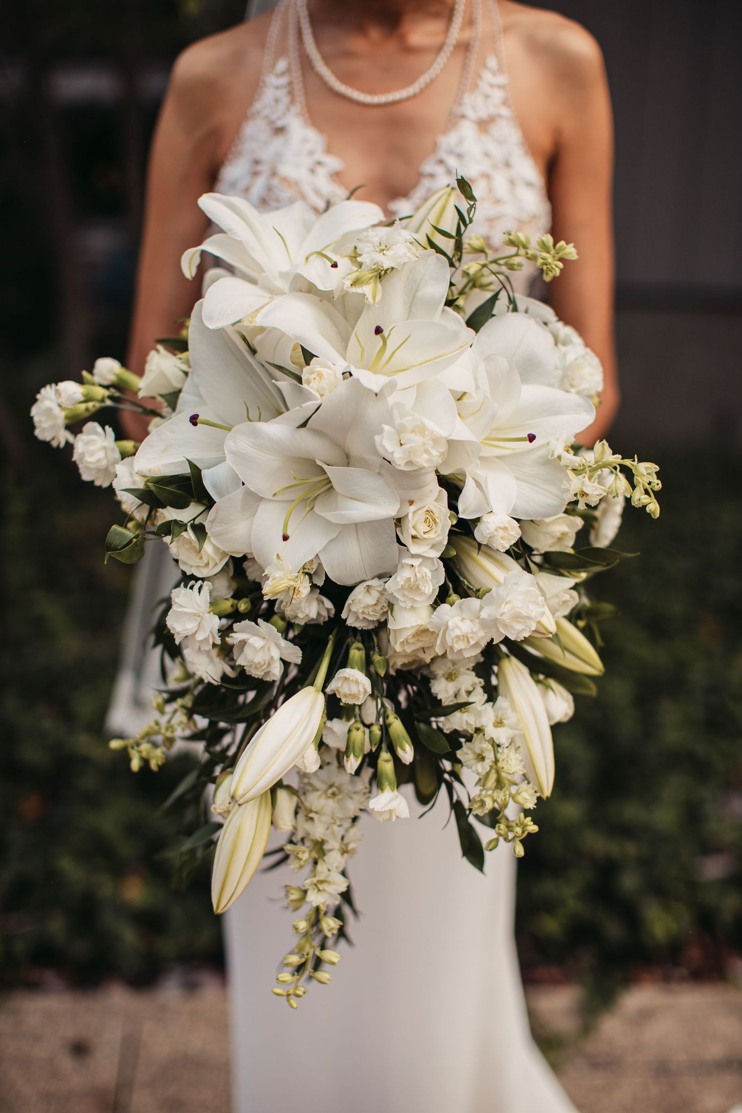 A cascading white wedding bouquet featuring elegant lilies, roses and delicate greenery against a wedding dress.