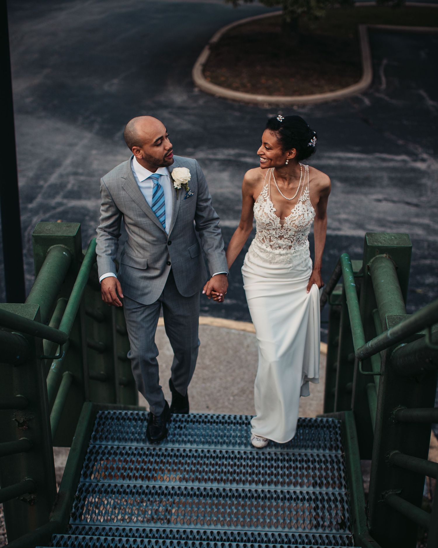 A happy couple wearing wedding attire walk together down green industrial stairs against a dark background.