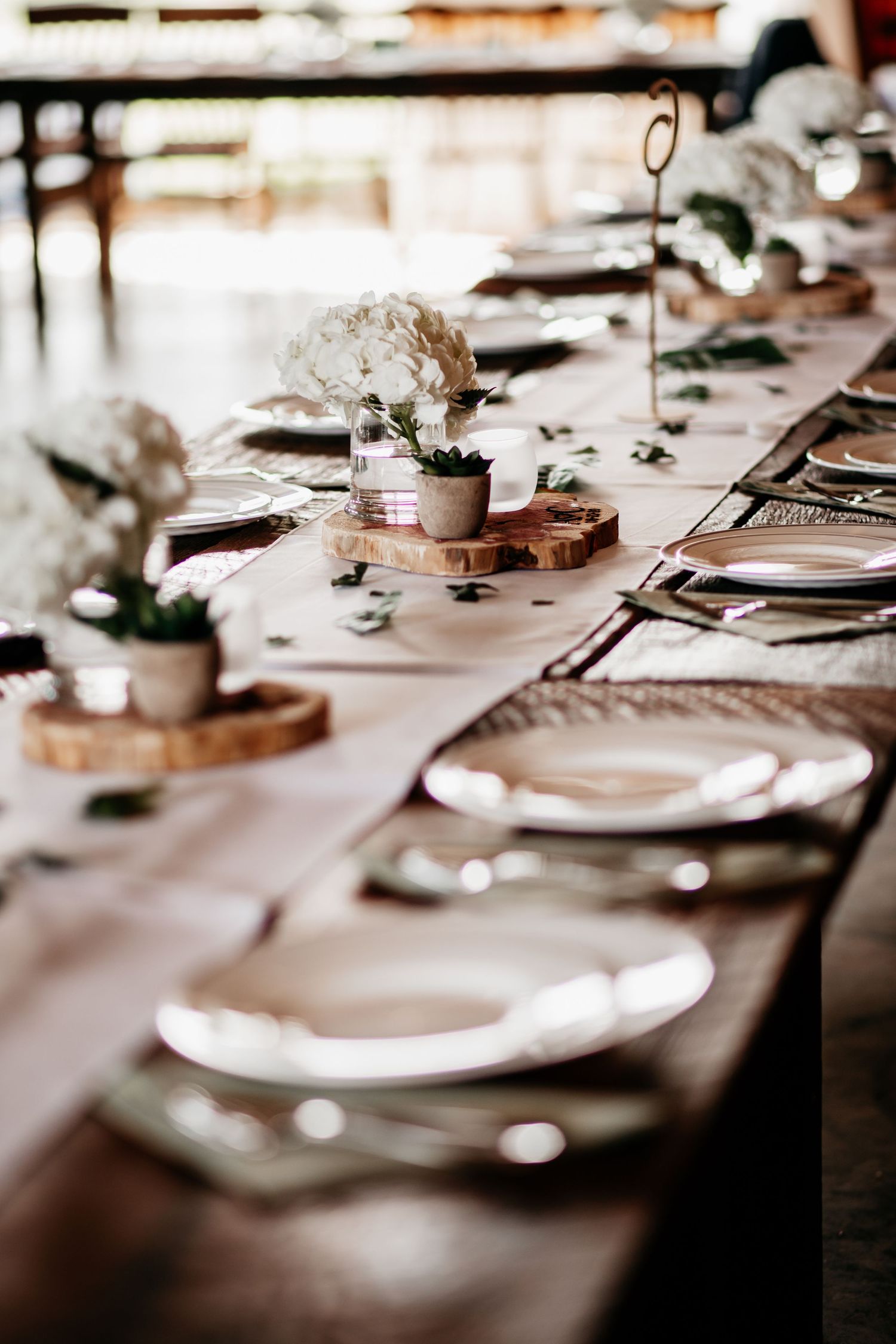Elegant wedding table setting featuring rustic wooden centerpieces, white plates, and delicate floral arrangements on a light tablecloth.
