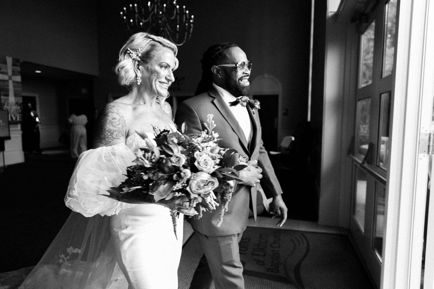 Black and white candid wedding photo capturing joyous moment with couple walking together while carrying floral bouquet.