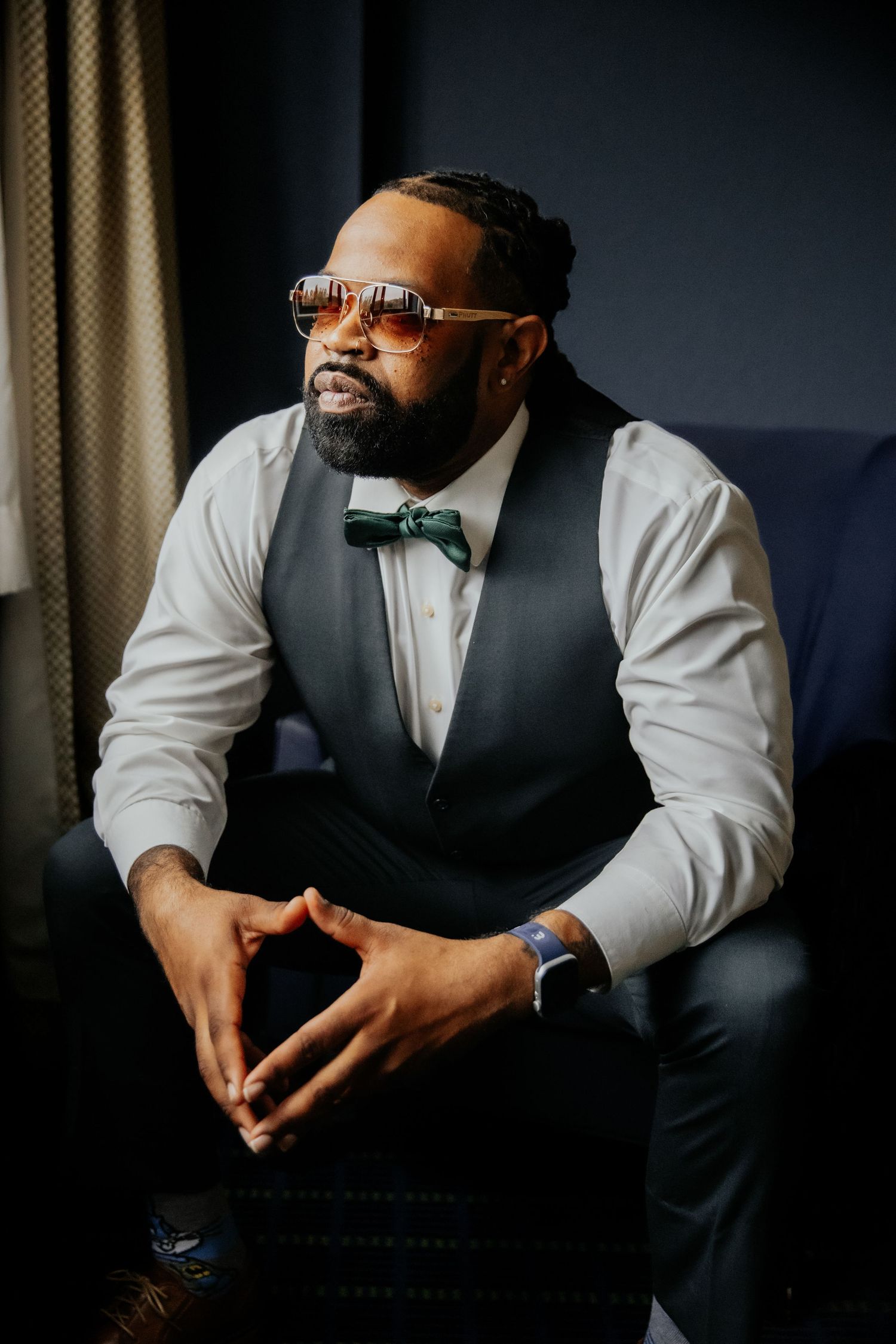 A well-dressed person in a white dress shirt, gray vest and bow tie sits contemplatively on a dark couch.