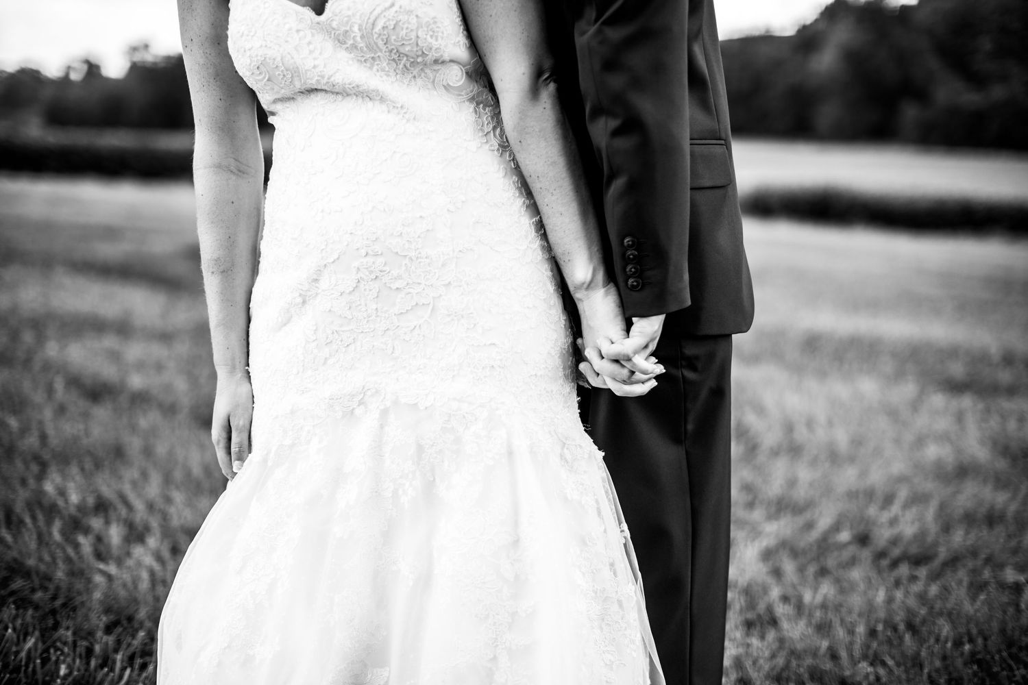 Black and white photo showing a bride and groom holding hands while standing together in a grassy field.
