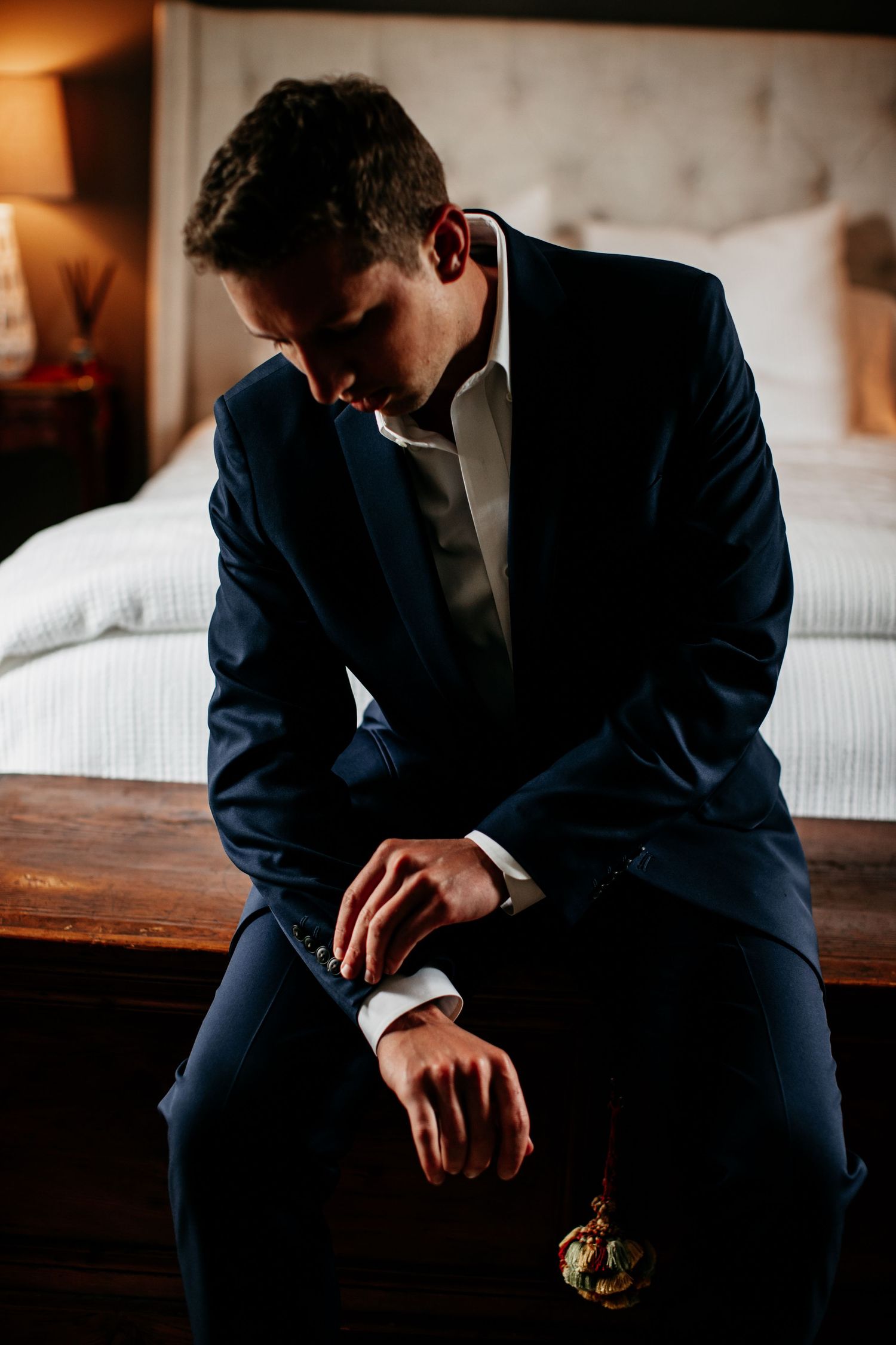 Silhouetted groom in navy suit sitting thoughtfully on bed edge in warm hotel room lighting.