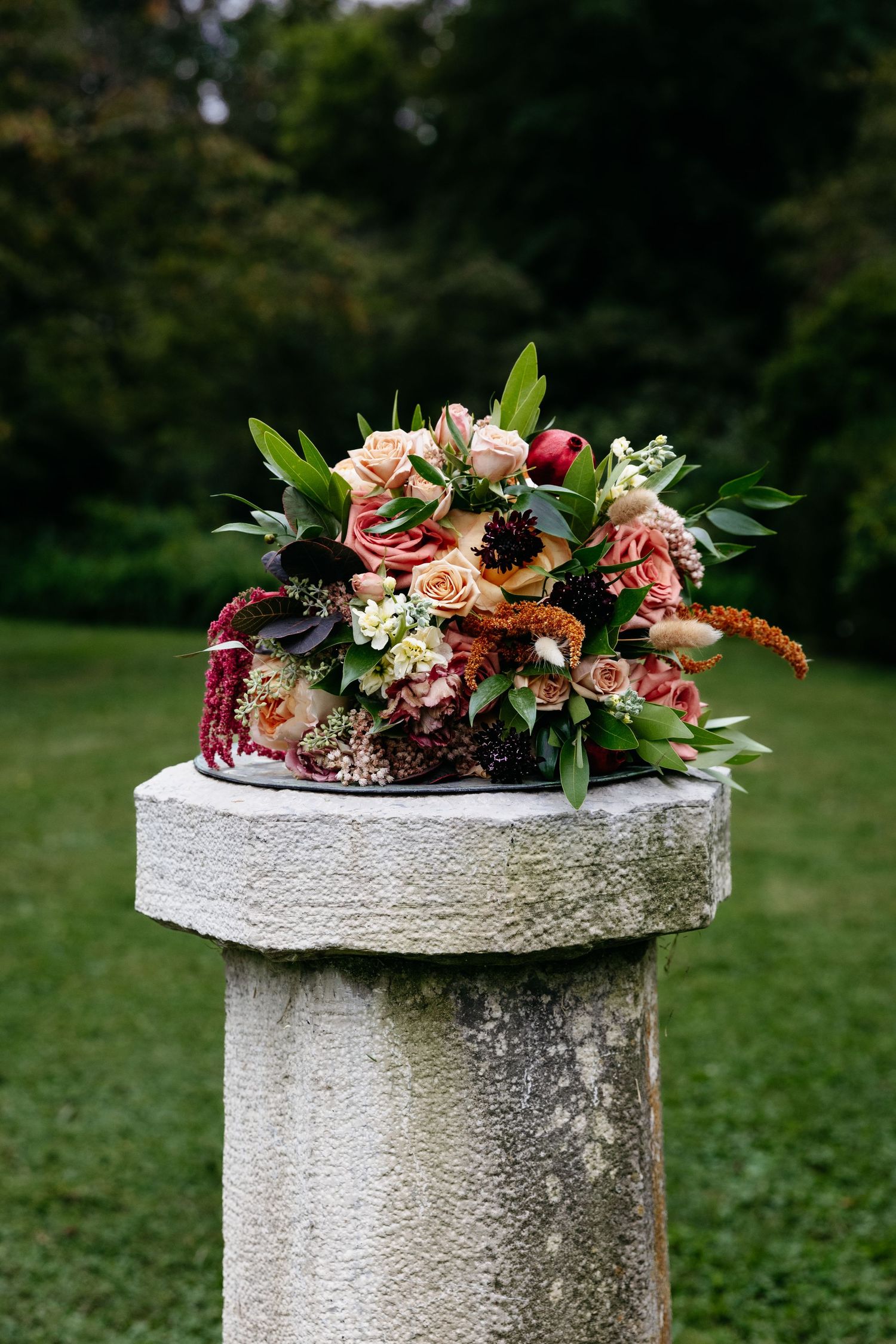 A beautiful wedding bouquet with pink and burgundy roses rests on a white stone pillar in a garden setting.