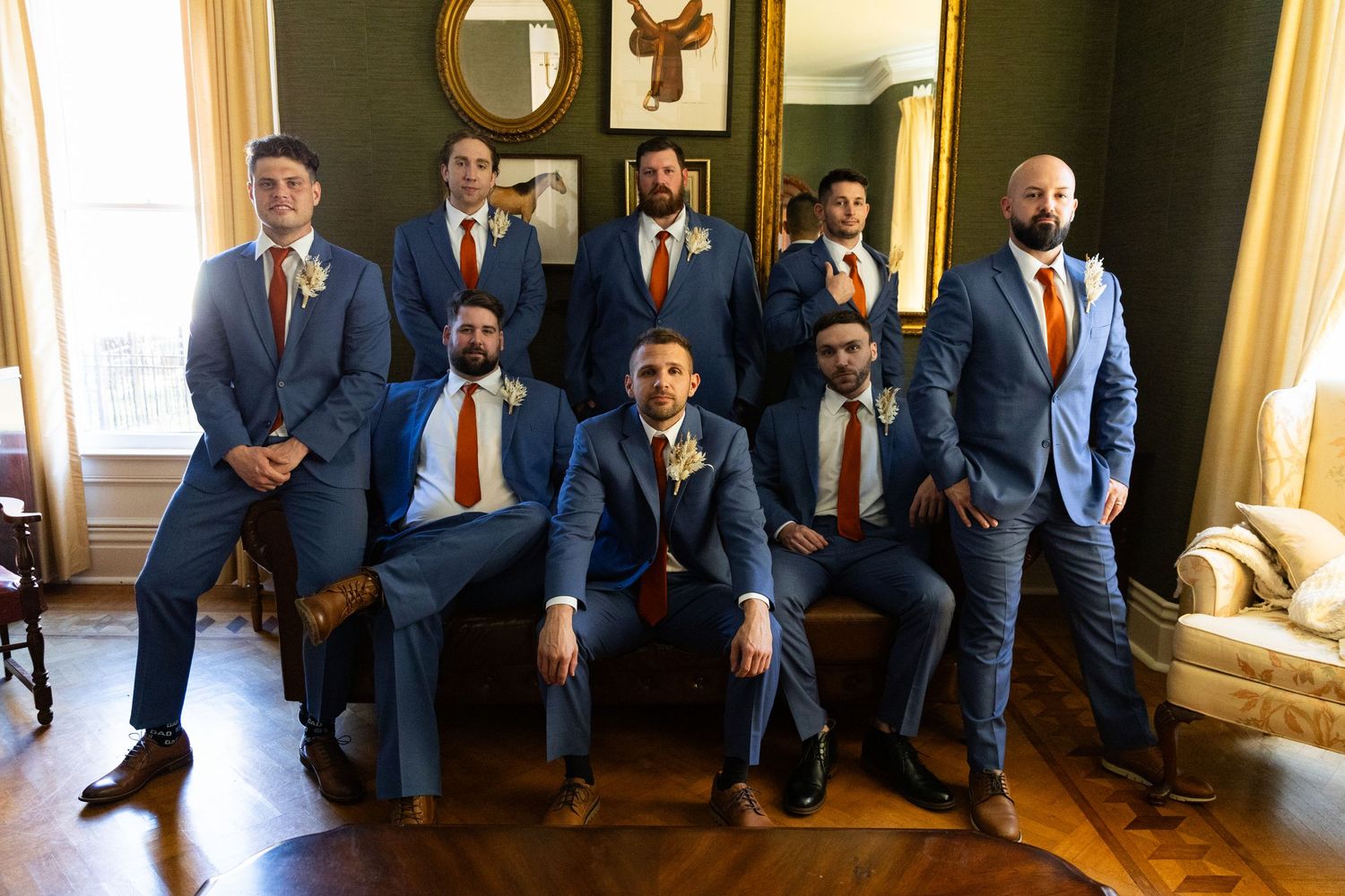 Wedding party in matching blue suits and red ties pose together in an elegant room with ornate gold mirror.