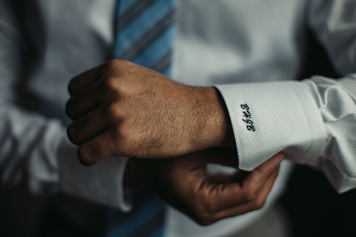 Hands adjust a white dress shirt cuff with pinstripes and blue tie visible in the background.