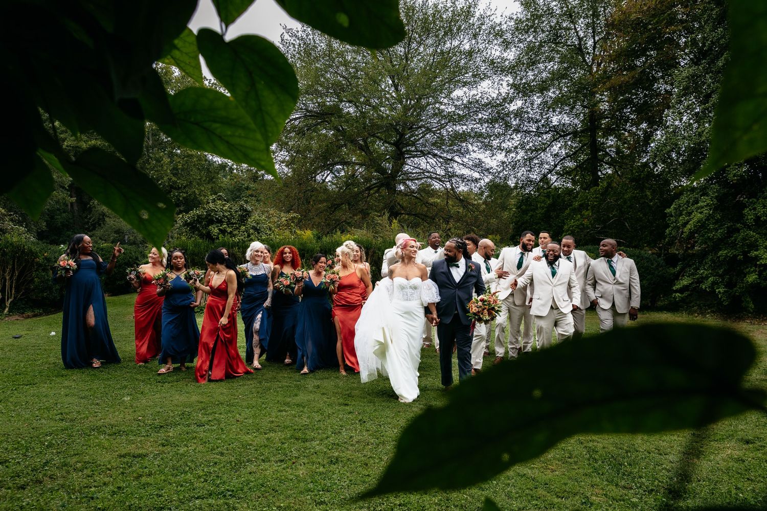 A wedding party in colorful formal attire poses together on a lush green lawn surrounded by trees.
