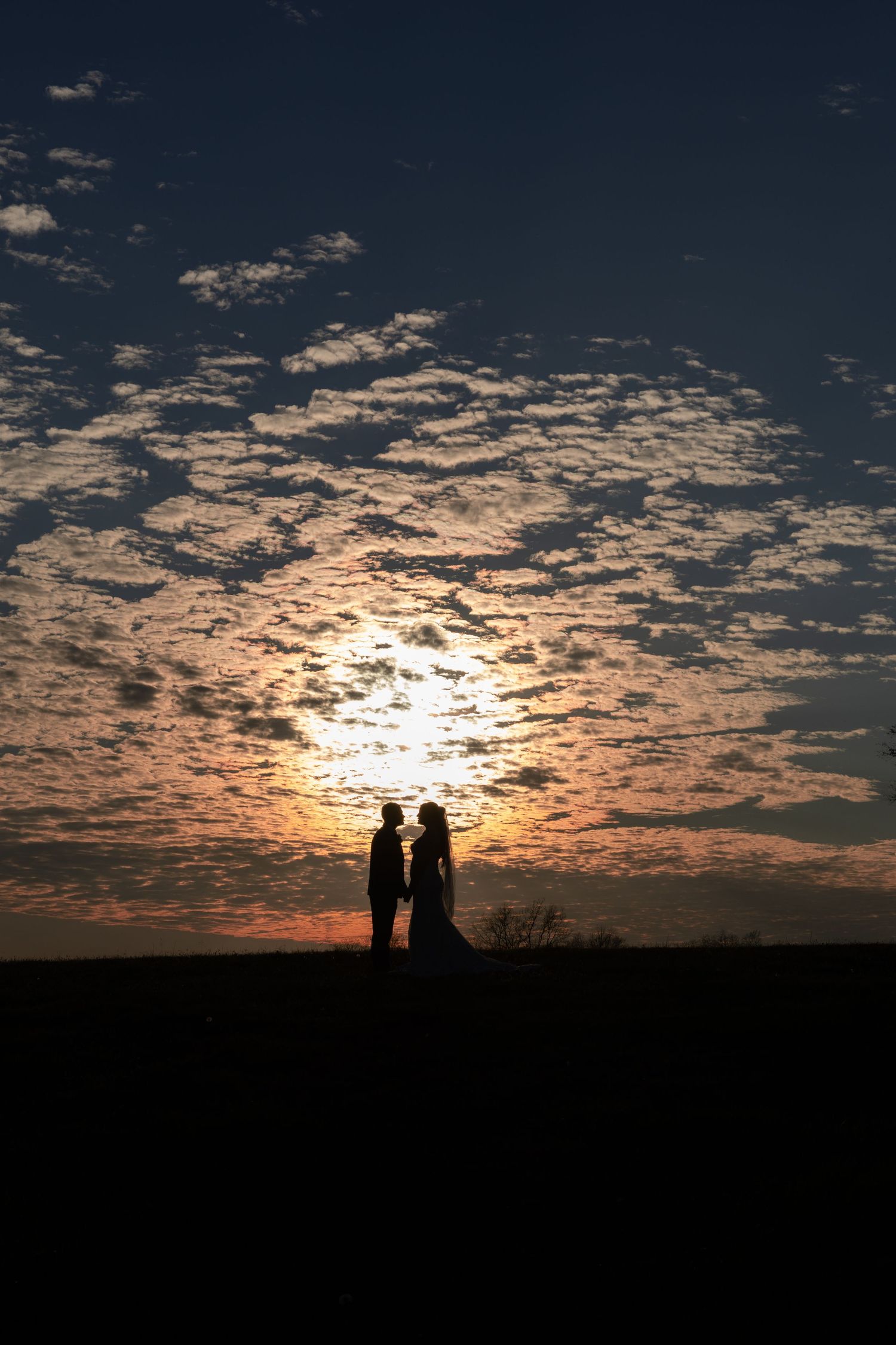 Two silhouettes stand together on a hill against a dramatic sunset sky with scattered clouds creating a golden glow.
