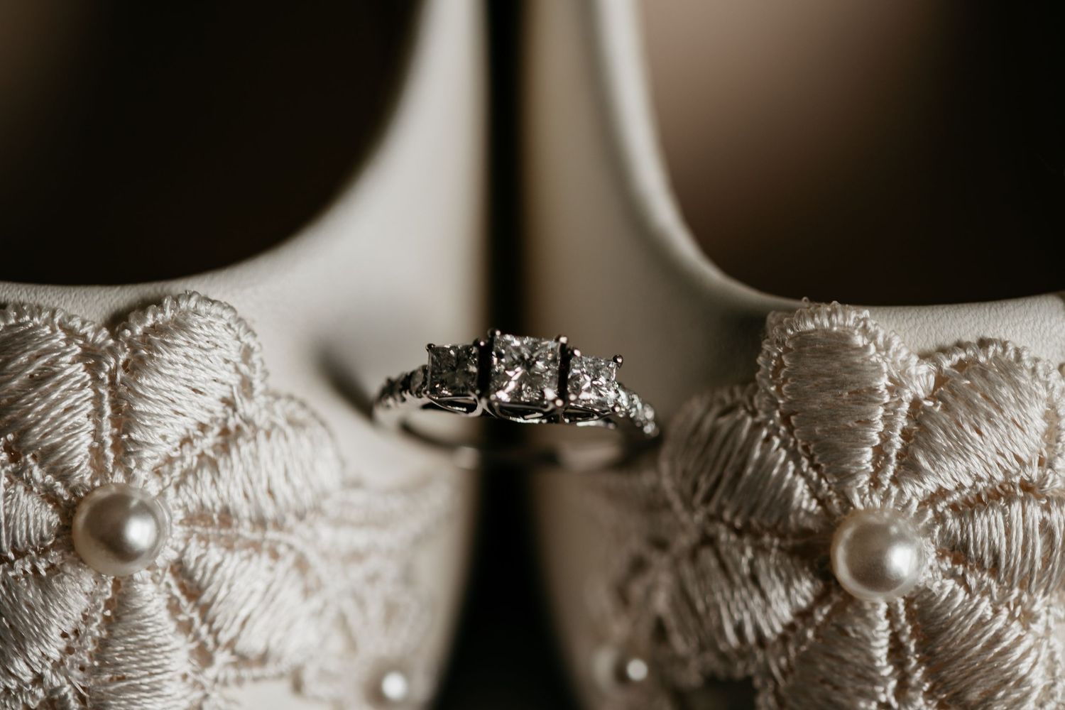 A diamond engagement ring displayed between two white lace bridal shoes with pearl details.