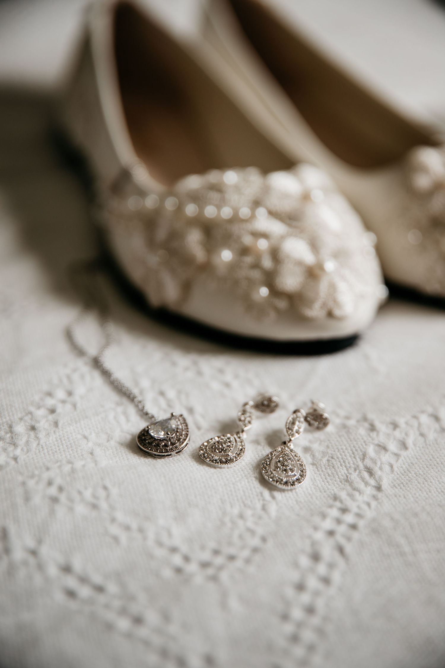 Vintage-inspired bridal shoes and crystal jewelry set displayed on delicate white lace fabric.