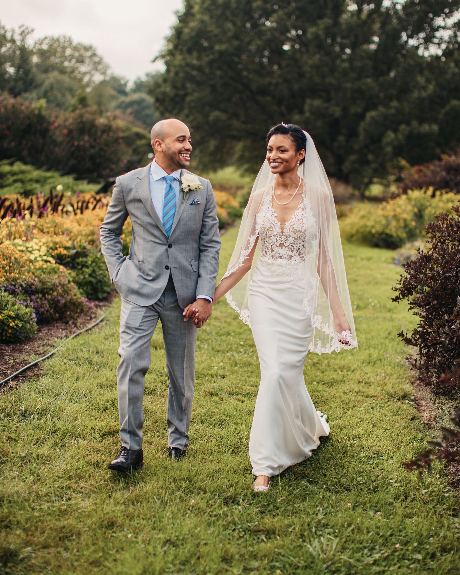 Happy newlyweds hold hands walking together outdoors, with bride in fitted lace gown and groom in light gray suit.