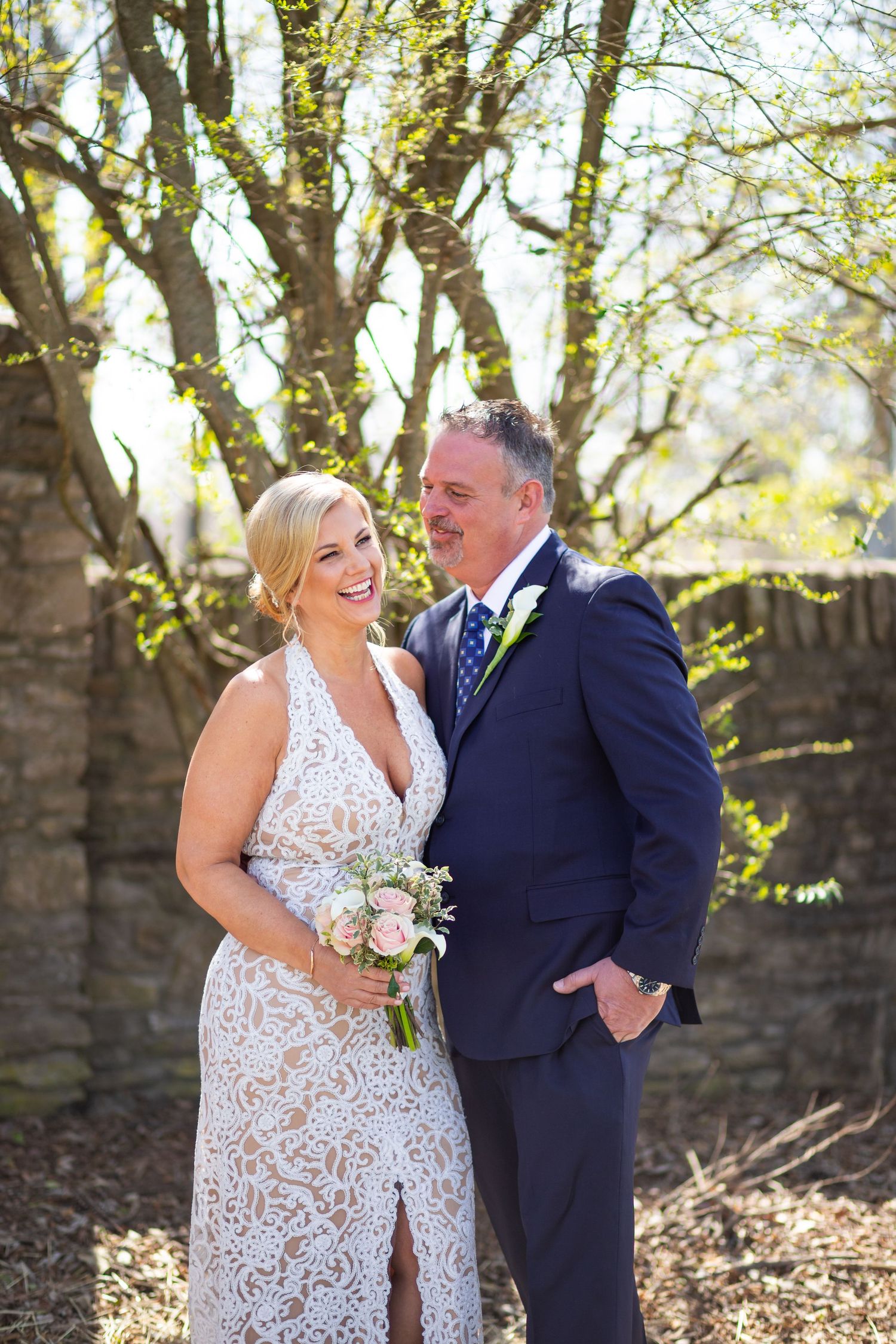 A couple in wedding attire shares a joyful moment among sunlit trees, with the bride holding a white floral bouquet.