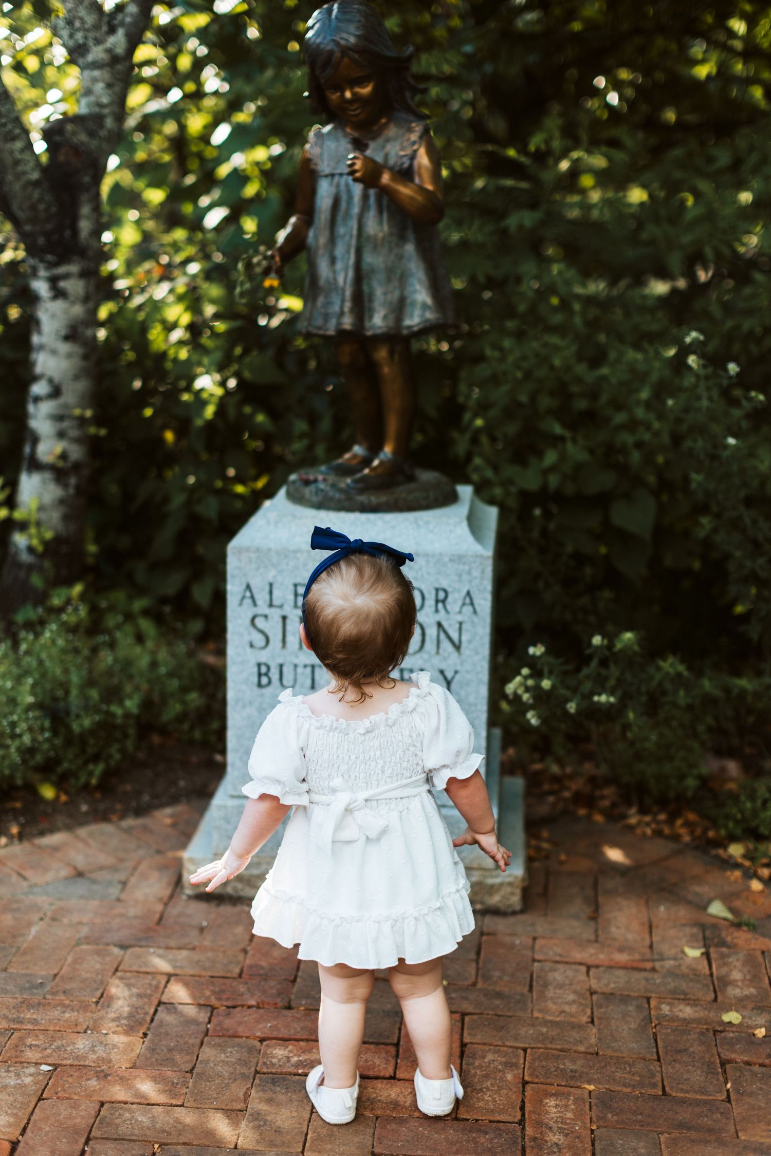 Small child in white dress looking up at bronze statue on pedestal in garden.